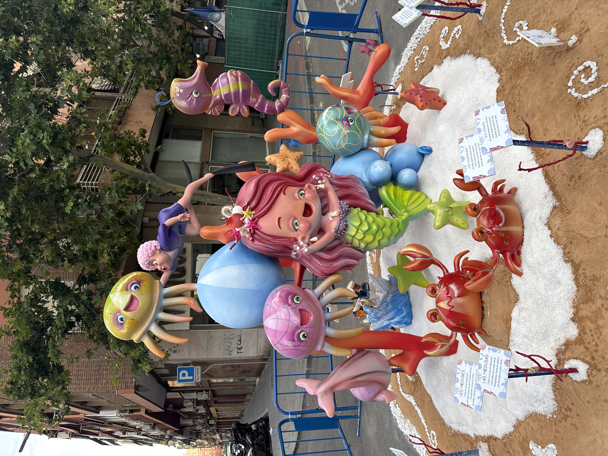An outdoor street scene features a large, colorful sculpture depicting an underwater theme. The sculpture is arranged on a sandy and white granular base, resembling a beach. Dominating the center is a mermaid character with vibrant pink hair and a green tail, holding small bubbles. Around her, various sea creatures are represented: a large yellow jellyfish with a smiling face, a pink jellyfish, an orange and purple seahorse, a pink dolphin, and two red crabs. Other elements include a large blue sphere, orange coral-like structures, and starfish. Several small informational signs with text and illustrations are placed around the base. In the background, there are buildings with balconies, trees with green foliage, parked motorcycles, and blue safety barriers. The sky is visible above the trees.