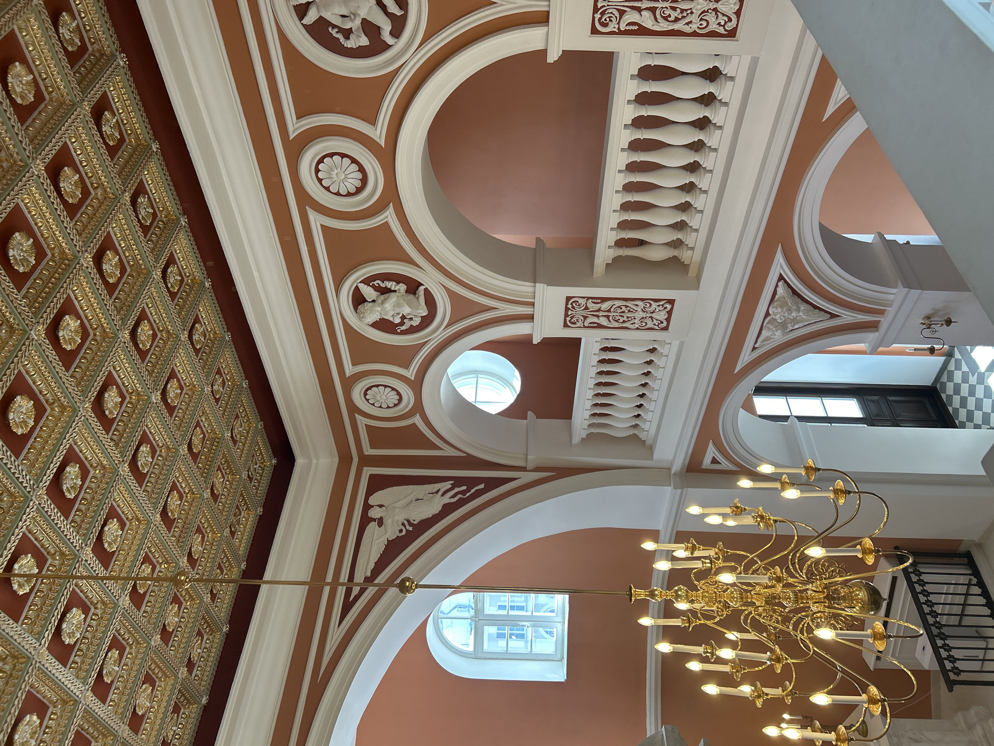 The image depicts an opulent interior with an ornate ceiling, a grand chandelier, and arched walkways with balustrades. The ceiling is a symphony of gold and deep red, featuring intricate geometric patterns and floral motifs. A large, multi-tiered brass chandelier with numerous candle-shaped lights hangs prominently, casting a warm glow.
The walls are painted a warm terracotta color and adorned with white stucco reliefs of cherubic figures, floral medallions, and winged beings. Arched openings lead to other parts of the building, and a white balustrade with decorative spindles lines a balcony overlooking a lower level. A round-topped window with white mullions is visible on the left, and another similar window is partially seen on the right. The flooring on the lower level, visible through an archway, is a checkered pattern of black and white tiles.
The provided context suggests the location is in Добасненскі сельскі Савет, Belarus. The scene appears to be a grand staircase or atrium within a historic building, such as a palace, museum, or a well-preserved public space. There are no people visible in the image. The overall impression is one of historical grandeur and meticulous craftsmanship.