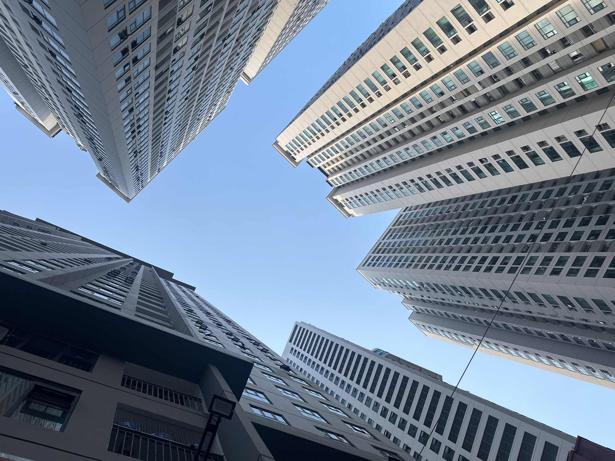 The image is a low-angle shot capturing five tall, modern buildings against a clear, blue sky. The buildings are primarily white and gray, with repeating patterns of windows across their facades. The perspective gives the impression of the buildings towering over the viewer, converging towards the center of the frame. The location is likely in a dense urban area, possibly Bac Nha Trang Ward, Vietnam, given the high concentration of high-rise buildings. No people are visible in the image.