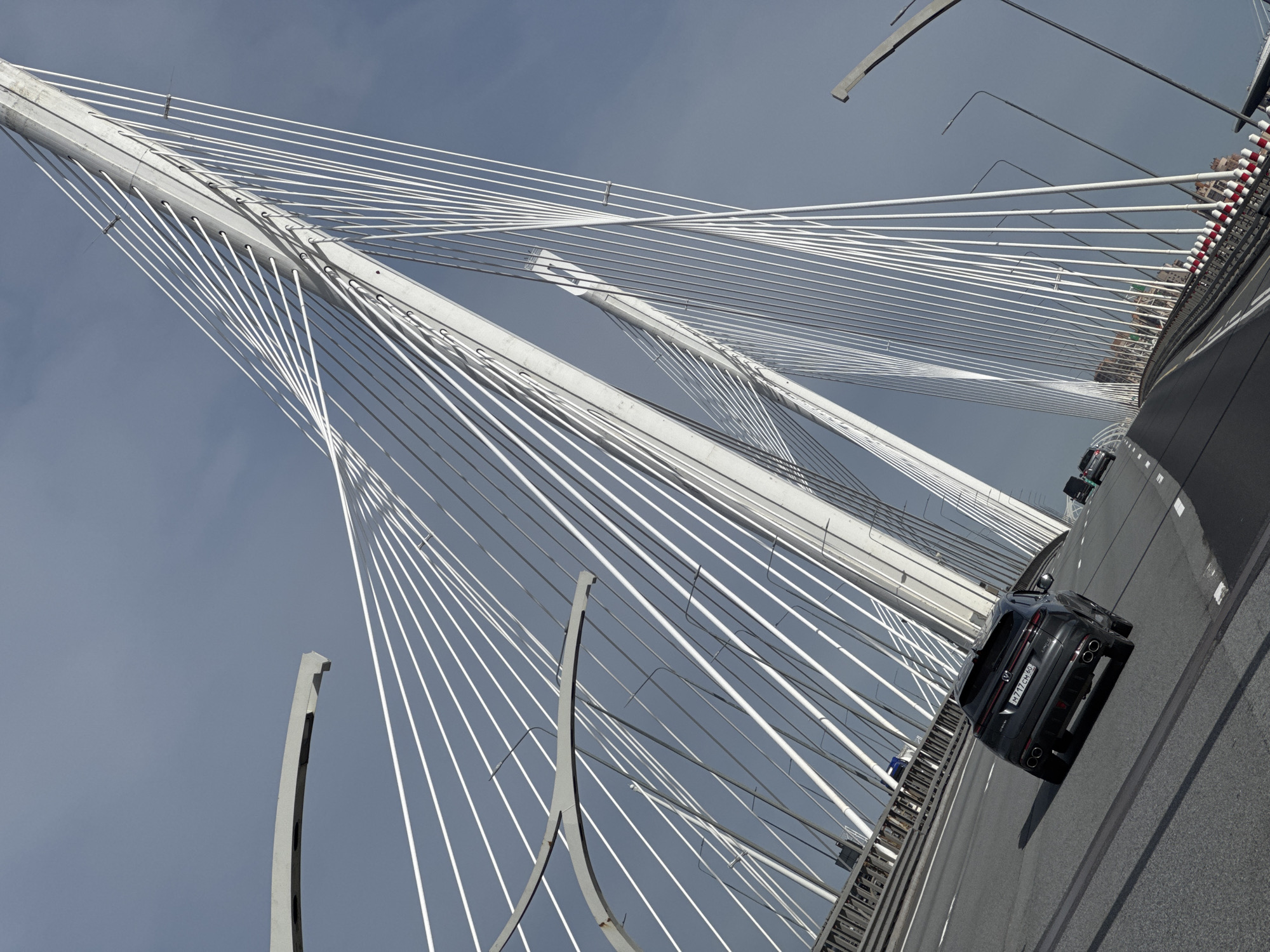 This image captures a low-angle, tilted perspective of a modern cable-stayed bridge against an overcast sky. The primary subjects are the stark white tension cables and the large, light gray concrete pylons from which they radiate, creating a complex, geometric pattern across the frame.
A dark-colored SUV is visible on the bridge deck, traveling away from the viewer. Its rear bumper features quad exhaust pipes, and a Russian license plate is clearly visible, displaying the text "M717CH 68". The bridge's roadway has visible white lane markings, and a barrier with red and white striped sections can be seen along the right edge of the road. In the distance, other faint vehicles and what appear to be urban structures are discernable.
The scene depicts a daytime setting with a gray, cloudy sky, suggesting overcast weather conditions. The architectural design of the bridge elements emphasizes its modern engineering. Based on the provided context, the location is Saint Petersburg, Russia, with the license plate region code (68) indicating Tambov Oblast, a common sight for vehicles from other regions in major cities. There are no discernible people or explicit events beyond routine traffic on the bridge.