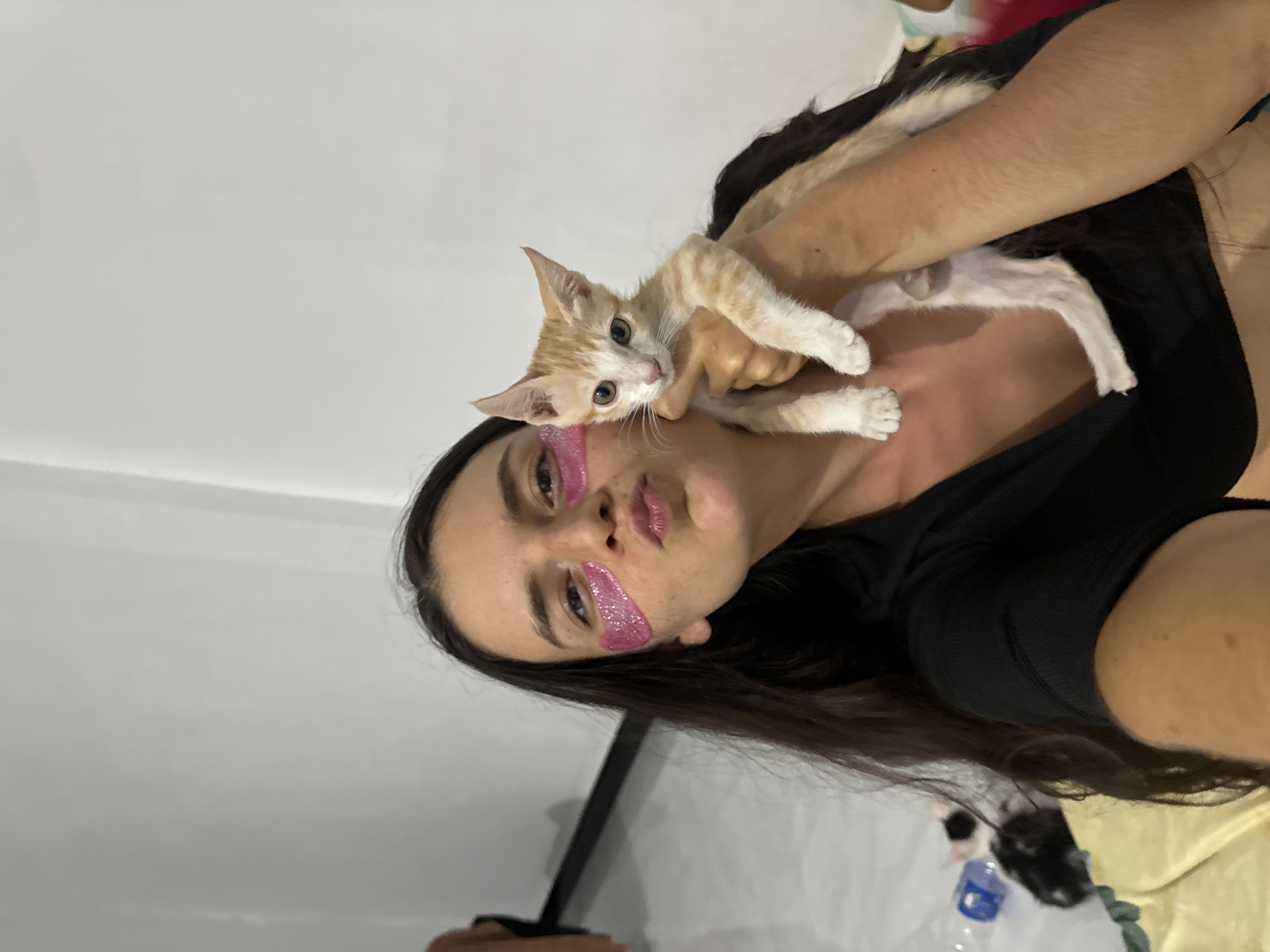 A young woman with long, dark hair is taking a selfie, holding an orange and white kitten on her shoulder. She is wearing a black top. She has pink glittery eye patches on under her eyes and pink glitter on her lips. The background is a plain white wall. The scene is indoors, possibly a bedroom or living area. The lighting appears to be artificial, suggesting it could be nighttime or an overcast day. The woman's expression is relaxed and affectionate as she interacts with the kitten. The kitten looks curious and is looking directly at the camera.
