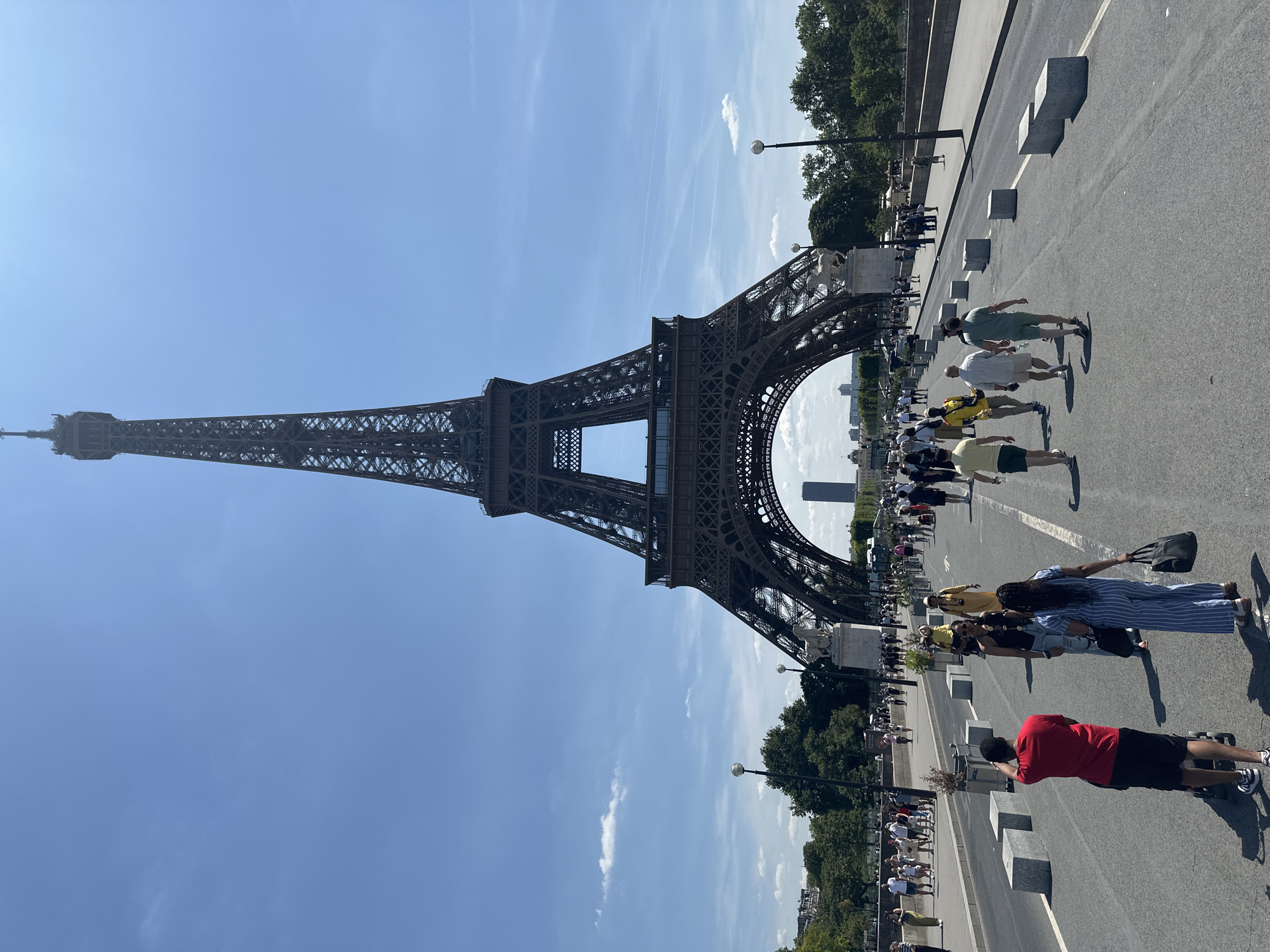 A wide-angle, eye-level shot captures the Eiffel Tower in Paris, France, on a clear, sunny day. The iron lattice structure of the iconic monument dominates the upper portion of the frame, with its base spanning the width of the image. In the foreground, a paved road is filled with people walking in various directions, some appearing to be tourists. Several concrete barriers are visible along the sides of the road. The atmosphere appears to be that of a busy tourist destination.