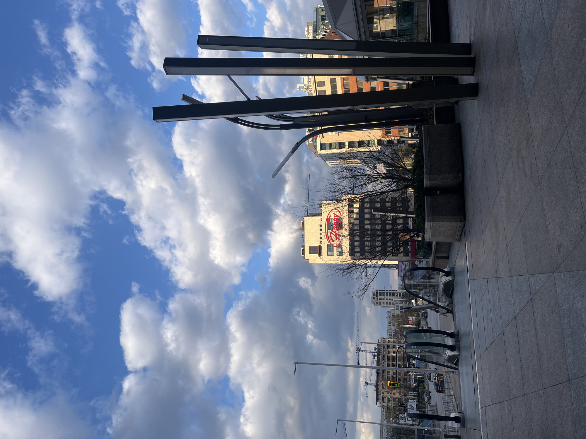 The image shows a view of Sofia, Bulgaria, likely taken from a street-level perspective. The sky dominates the left portion of the frame, featuring a mix of blue sky and large, puffy white clouds. A modern building with a red "Mall of Sofia" sign is visible in the mid-ground. In the foreground, there are several dark, vertical metal structures that appear to be part of a building or urban design element. The right side of the image shows a paved area, possibly a sidewalk or plaza, with some greenery in planters. Escallators can be seen moving from street level up to the plaza, suggesting the photograph was taken at the entrance of a shopping area. The photograph is notable for its depiction of urban architecture against a dynamic sky. The image includes construction cranes in the distance, suggesting ongoing building activity.