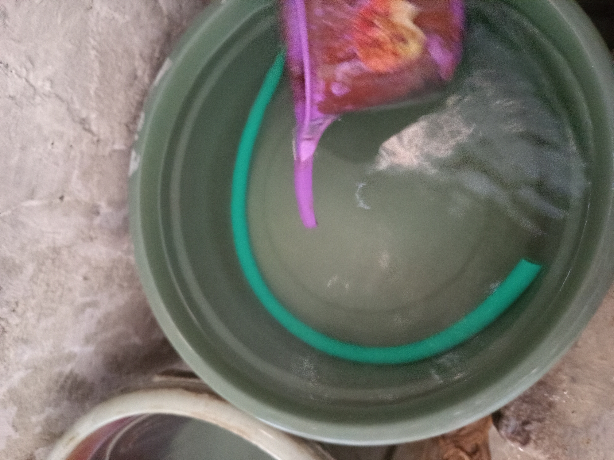 A green plastic tub filled with water sits on a textured surface, possibly concrete. A green hose is coiled inside the tub. A purple watering can with a floral design is partially submerged in the water. Another white tub is visible to the left, also containing water. The scene appears to be an outdoor setting, likely in a backyard or alleyway in the city of Tebel, Indonesia.