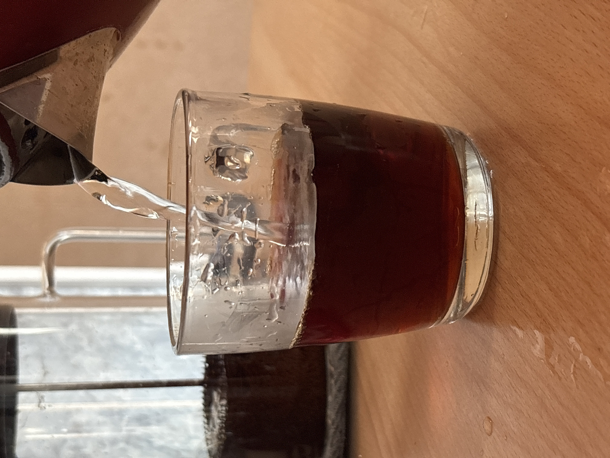 A clear glass filled with ice and a dark liquid, likely soda or iced tea, is being poured into from a metal pitcher. The glass sits on a light brown wooden surface. A metal rack or dish drainer is visible in the background. The image does not suggest any connection to Chaikovsky, Russia.