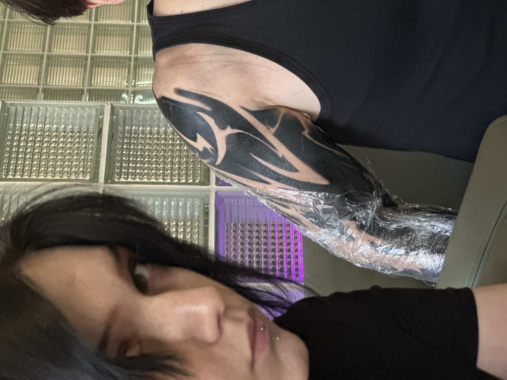 This media file captures an indoor scene, likely a tattoo studio in Moscow, Russia. The primary focus is on a person's left arm and upper torso, wearing a black tank top, positioned on the right side of the image. The arm features a large, fresh tribal-style tattoo composed of solid black shapes and negative space patterns. The tattooed area is partially covered in clear plastic wrap, indicating it is a recent tattoo or part of the healing process.
In the foreground, on the left side, is a second individual. This person has dark hair and a neutral expression, looking towards the camera. They are wearing a black t-shirt and have a nose piercing visible on their philtrum.
The background consists of a wall constructed from glass block tiles. Some of these blocks appear clear or frosted, while others in the lower left corner emit a distinct purple light, possibly from an accent light source behind them. The overall lighting suggests an indoor setting, likely with artificial illumination. No visible text is present in the image.