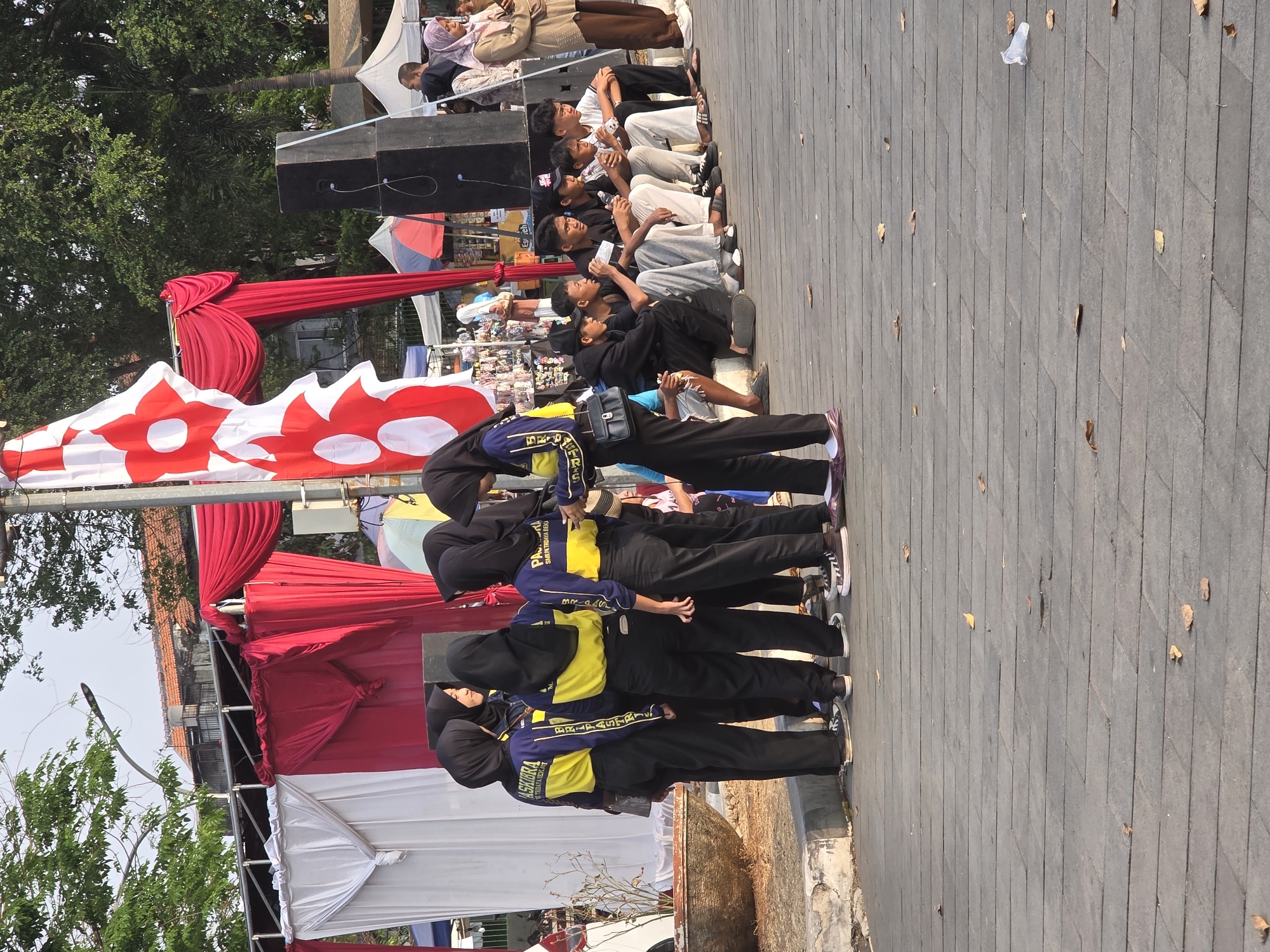 In Tambun, Indonesia, a group of six young women wearing black hijabs and matching blue and yellow uniforms stand clustered together on a paved outdoor area. One woman on the far right is turned away, while the others face inwards, engaged in conversation. A large red and white flag is positioned behind them, draped from a pole. To their right, a group of young men are seated on the ground, some holding drinks. In the background, tents and market stalls are visible, along with trees and buildings. The ground is paved with grey rectangular stones, and fallen leaves are scattered across the surface. The scene suggests an outdoor gathering or event.