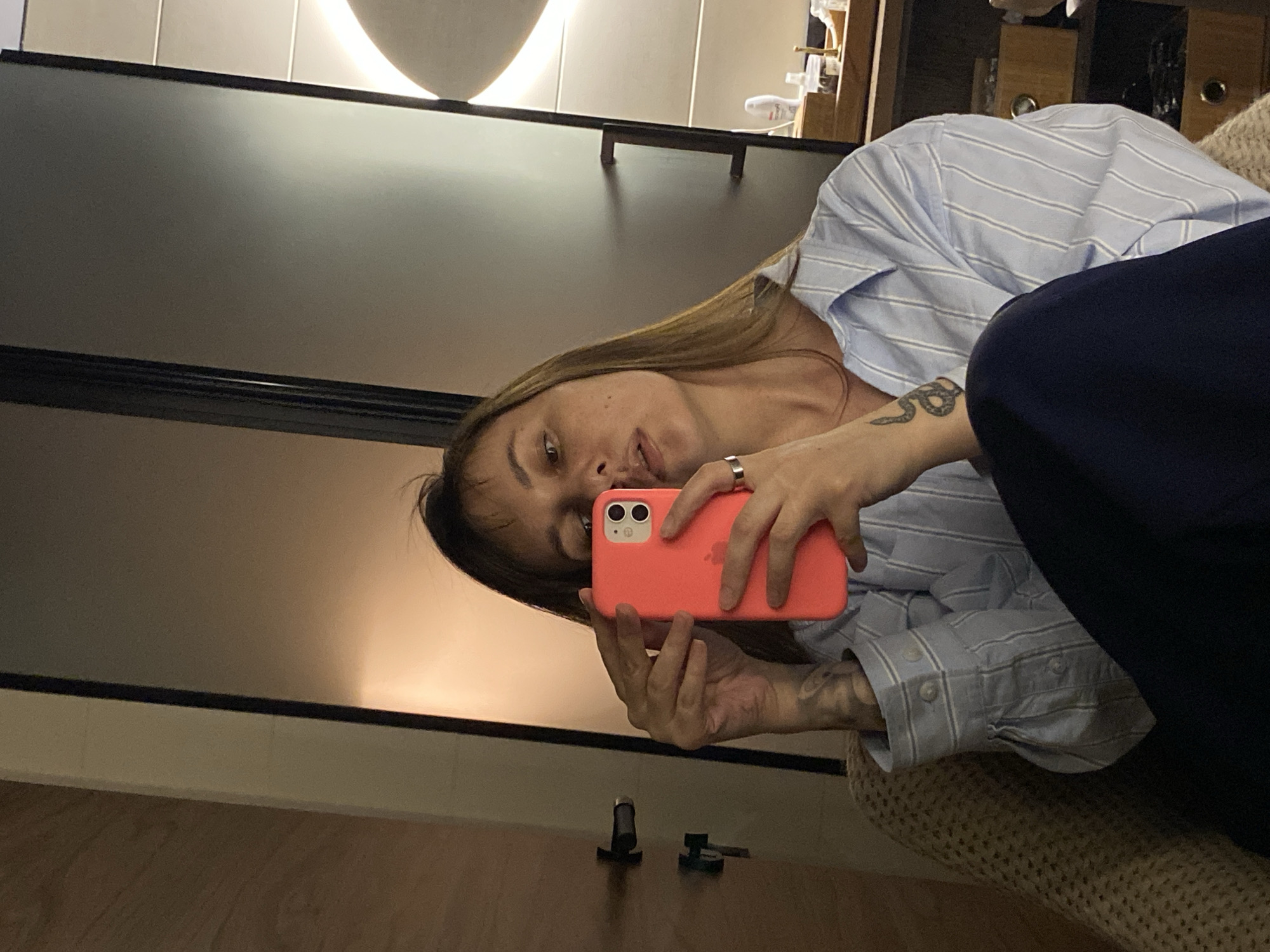A woman is taking a selfie with a coral-colored phone. She is wearing a striped blue shirt and dark blue pants. A snake tattoo is visible on her left forearm. She is sitting on a textured beige chair in what appears to be an indoor setting with a mirror and wooden floor. The precise location within Votkinsk, Russia, cannot be determined from the image.