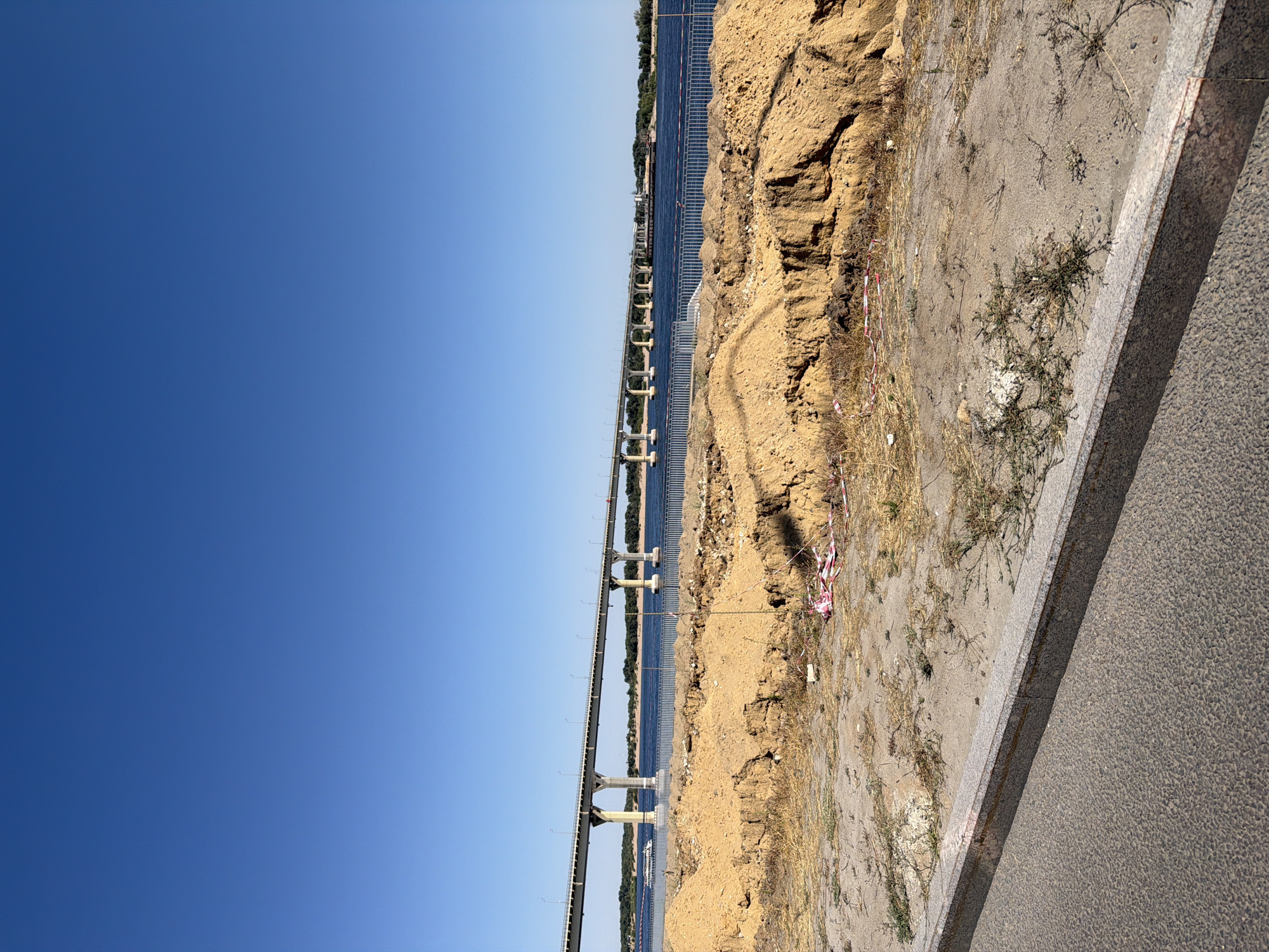 The image shows a wide, calm river under a clear blue sky. A long, multi-span bridge stretches across the river, with its concrete piers supporting the roadway. A white passenger boat is visible on the water near the bridge. In the foreground, there is an area of disturbed earth, with piles of sand and some scattered debris, including red and white caution tape. A concrete curb separates this area from an asphalt surface in the immediate foreground. The scene appears to be an outdoor construction or renovation site near the river. The bridge in the image is the Volga Bridge in Volgograd, Russia, which is a significant transportation artery over the Volga River.