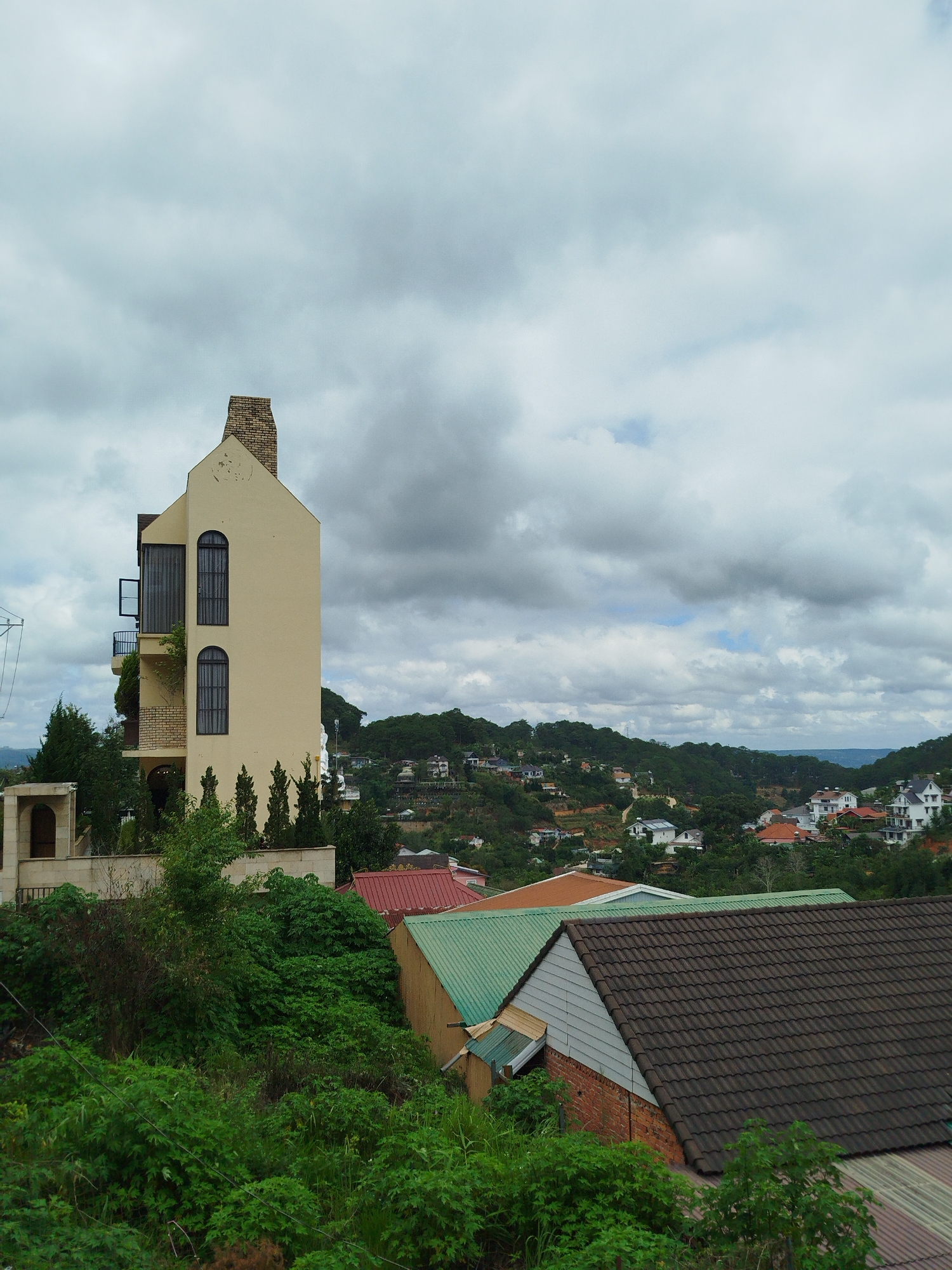 The image captures a panoramic view of a residential area in Da Lat, Vietnam, characterized by a hilly landscape and a mix of architectural styles. In the foreground, lush green vegetation dominates, with dense bushes and trees creating a natural barrier. Several rooftops are visible, including a building with a dark brown tiled roof in the lower right, another with a green metal roof, and a reddish-brown tiled roof further back.
A prominent structure on the left is a tall, slender, pale yellow house with a steep, gabled roof and a brick chimney. This house features arched windows with dark grates and a small balcony on the side. The surrounding terrain is sloped, suggesting the location is in a hillside district.
In the middle ground and background, the landscape unfolds into a sprawling hillside covered with numerous houses, many of which appear to be white with red or brown roofs. These homes are nestled amongst dense evergreen trees, giving the impression of a verdant, forest-like environment. The houses are scattered across the slopes, with some roads or pathways visible winding through the community. The overall impression is of a moderately overcast day, with diffused light illuminating the scene and a cloudy sky overhead. The scene is devoid of any visible people or animals.