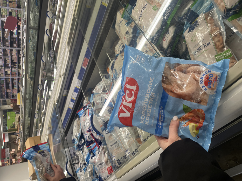 The image captures a close-up view inside a supermarket, specifically in the frozen food section. The main subject is a person's hand holding a light blue plastic bag of frozen shrimp from a display freezer. The hand is visible from the wrist down, clad in a dark sleeve, and shows fingernails painted a light, possibly white, color.
The bag prominently displays the brand name "VICI" in red lettering on a white background within a red rectangle. Below this, large Cyrillic text reads "КОРОЛЕВСКИЕ КРЕВЕТКИ" (Royal Shrimps). Further down, it specifies "в панцире с головой" (in shell with head) and "С ПРЯНОСТЯМИ" (with spices). The bag features an image of a cooked shrimp with a slice of lemon and a sprig of herb at the bottom right. A circular blue sticker on the bag contains more Cyrillic text, including "ЮЖНОЙ" (Southern) and "50/70 ШТУК" (50/70 pieces), indicating the size or count of the shrimp. The product inside the bag is visible through a clear window, showing frozen, pale pink shrimp. Frost and condensation are visible on the outside of the bag.
The background shows the interior of a refrigerated display case, filled with numerous other bags of frozen seafood, primarily shrimp. Many of these bags are white or clear plastic, and some display the text "КРЕВЕТКИ" (Shrimps) and numbers like "100/200" which likely denote shrimp size. Other partially visible English and German words like "SHRIMPS GARNELEN" also appear on some packaging. Blue price tags and product information strips are attached to the shelving inside the freezer. One price tag on a shelf above is partially visible showing "1405" and other numbers.
Beyond the freezer, the image's perspective is rotated, showing rows of shelves stocked with various products, typical of a supermarket aisle, reflected in the freezer's glass door. A pink ice cream bar-shaped sign or decoration is hanging in the distance in the reflected aisle. The overall lighting is bright and even, characteristic of an indoor retail environment.