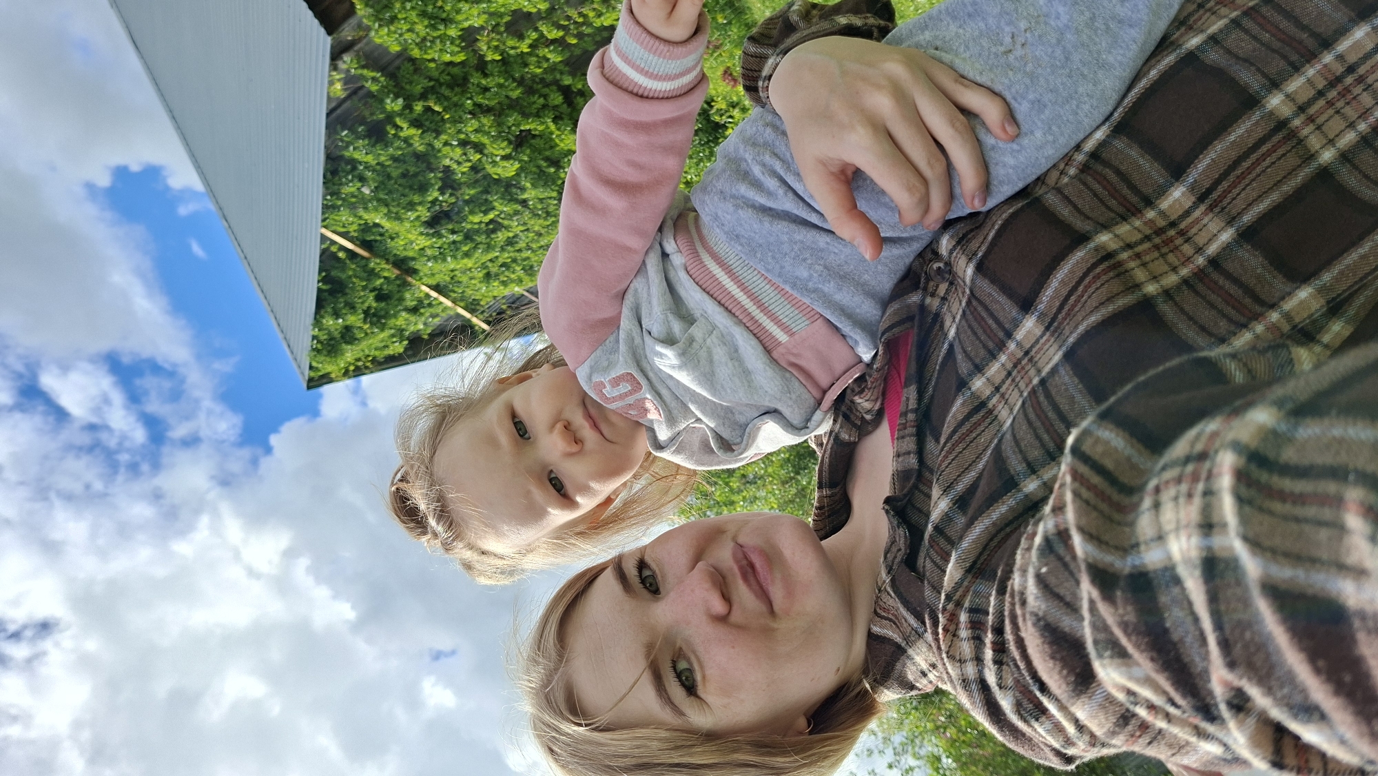 An outdoor selfie, rotated 90 degrees counter-clockwise, captures a woman and a young child in a bright, natural setting. The woman, with blonde hair and green eyes, is positioned in the lower right of the frame, looking directly at the camera with a soft, content expression. She wears a brown, tan, and red plaid shirt, with a hint of a pink garment visible underneath.
The young child, likely a girl with light blonde hair and green eyes, is held by the woman, appearing above her in the upper left of the frame. The child also looks towards the camera with a calm, curious expression. She is dressed in a grey long-sleeved top or hoodie featuring pink cuffs, a pink collar, and pink stripes near the chest.
The background, also rotated, shows a clear blue sky with fluffy white clouds dominating the left side of the image. On the right, lush green foliage, likely trees or bushes, is visible, along with part of a white building with a slanted roof. The lighting suggests it is daytime with bright, natural sunlight.
The overall atmosphere is casual and affectionate, capturing a shared moment between the two subjects. No visible text is present in the image. The scene takes place outdoors in Большой Букор, Russia, suggesting a rural or suburban environment.