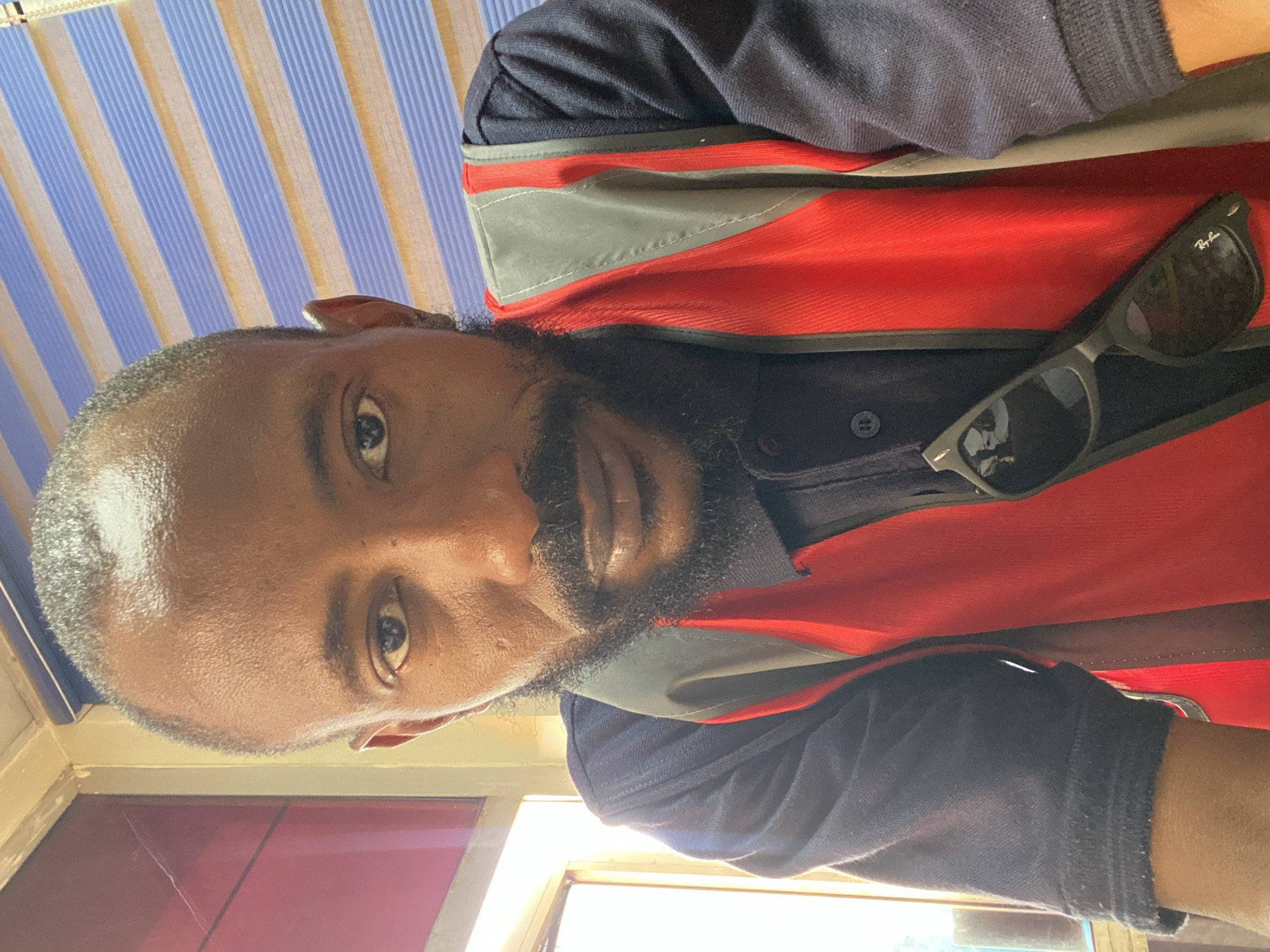 An adult man, likely of African descent, is featured in a close-up selfie, taken with a sideways orientation. He has dark skin, a short, well-groomed beard, and closely cropped dark hair. He looks directly at the camera with a slight, neutral smile.
He is dressed in a dark (possibly navy blue or black) collared polo shirt, worn under a red and grey jacket or vest. The jacket has distinct grey panels, possibly reflective, across the chest. A pair of black Wayfarer-style sunglasses, bearing the "Ray-Ban" logo on the right lens, is hooked onto the front of his top.
The background shows a blue and light tan striped awning or ceiling structure in the upper portion of the image. The lower part features a window through which bright daylight is visible, along with reddish-brown translucent panels or blinds. The bright natural light suggests the photo was taken during the daytime. The setting appears to be an indoor or covered outdoor area, possibly a casual establishment in Sabon Gari Nassarawa, Nigeria. The man is posing for the photo.