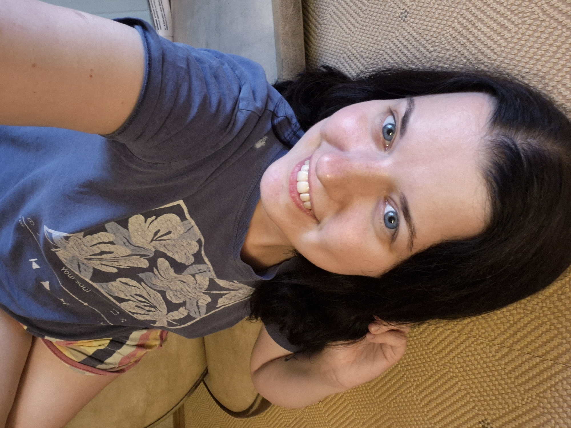 The image shows a close-up, selfie-style portrait of a young woman with dark hair and blue eyes. She is smiling and looking directly at the camera. She is wearing a dark blue t-shirt with a floral pattern and what appears to be colorful striped shorts. The background is a textured, yellowish-tan surface, possibly upholstery of a couch or chair. The lighting suggests an indoor setting, and the overall impression is casual and relaxed. No specific location within Saint Petersburg, Russia, can be determined from the image itself.