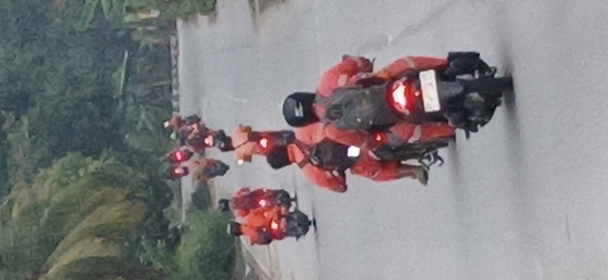 A convoy of motorcyclists wearing orange jumpsuits ride down a paved road flanked by dense foliage. The motorcyclists are viewed from behind, and many have backpacks. Their motorcycles have red taillights illuminated. The scene appears to be on a slightly overcast day. The location is the city of Tanjung Balai Karimun, Indonesia.