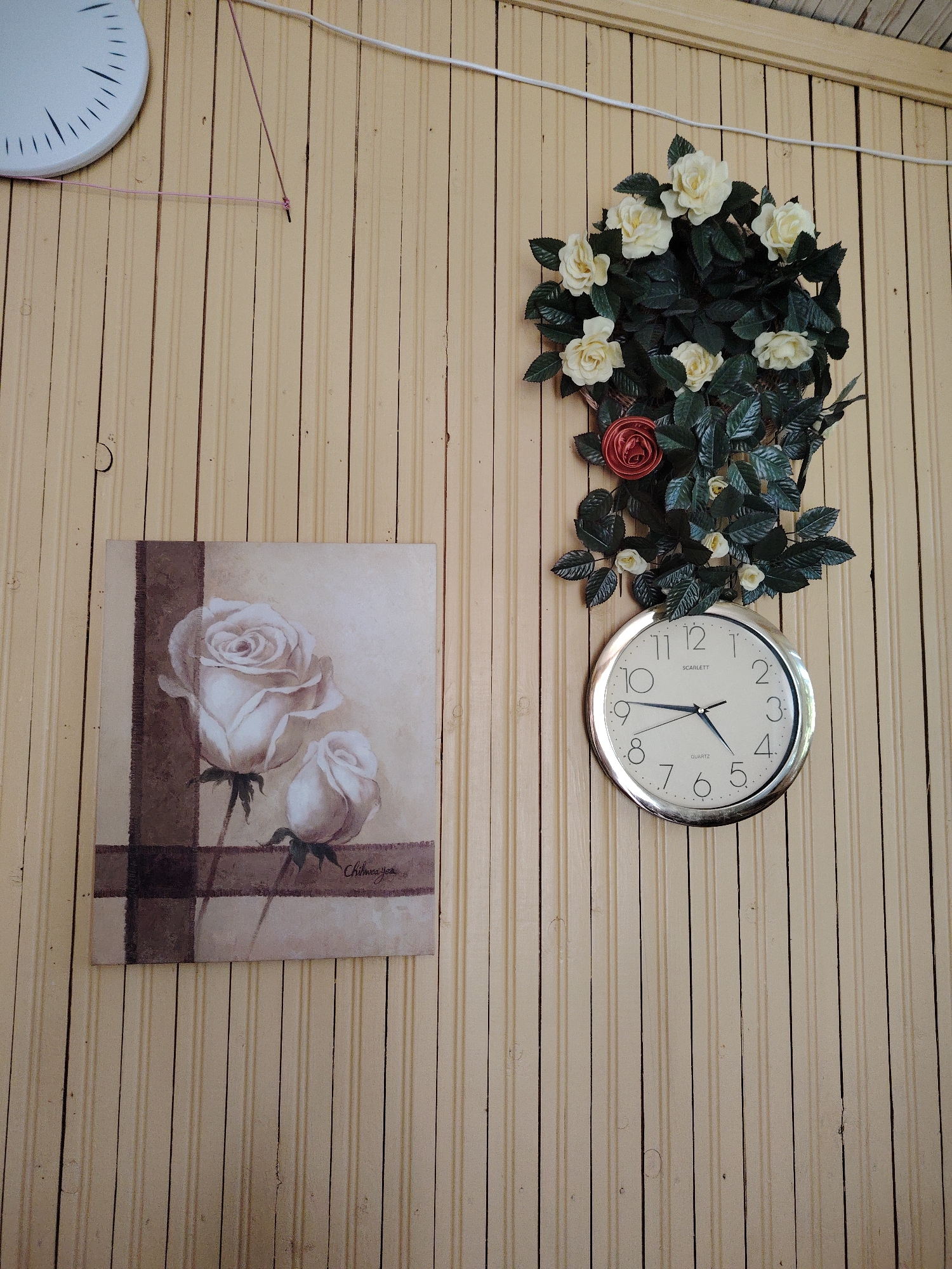 A close-up shot shows a wall with vertical pale yellow wooden paneling. On the left side of the wall, a white clock with black markings is partially visible. In the center of the image, a framed artwork depicts two white roses with brown accents. To the right of the artwork, a round clock with a silver frame hangs on the wall, with a wreath of green leaves and white and red roses above it. The clock shows the time as approximately 4:43. The overall scene appears to be indoors.