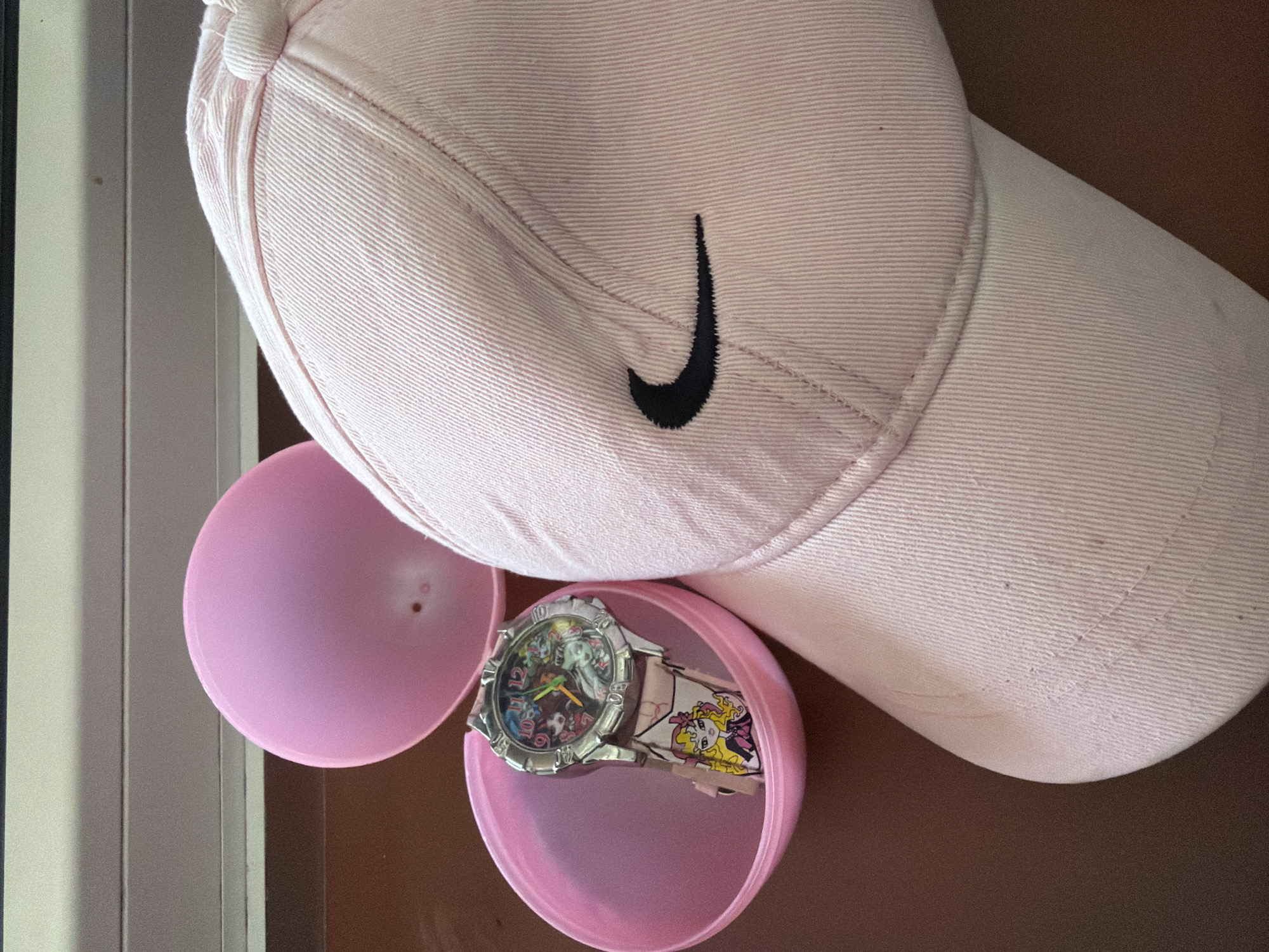 A pink Nike baseball cap is placed on a brown surface next to two halves of an open pink plastic egg. A doll-themed watch is inside one half of the egg. The scene appears to be indoors. There is no indication of the city administration of Qarağandı, Kazakhstan.