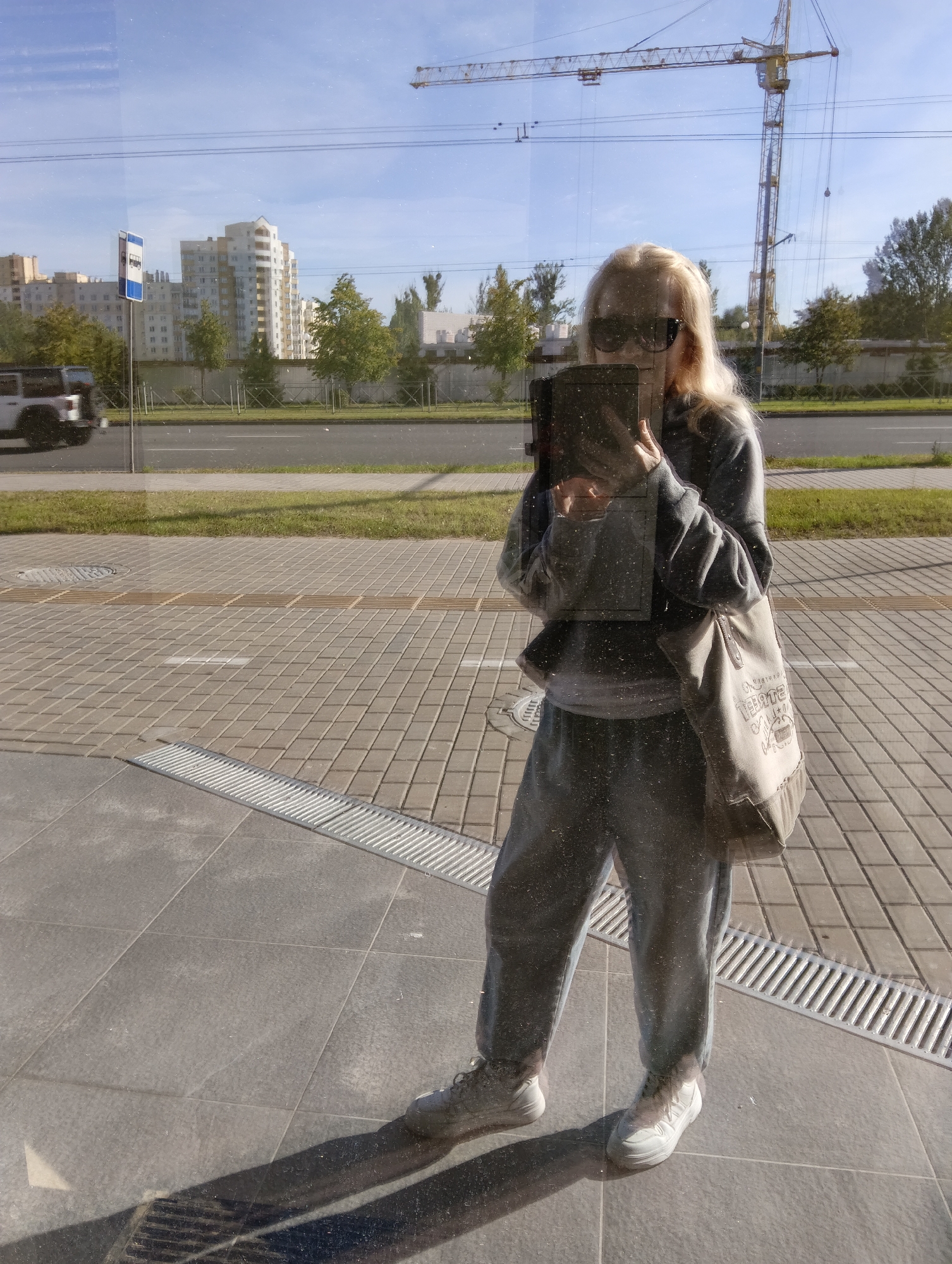 The image shows a full-body mirror reflection of a woman standing on a sidewalk in Hrodna, Belarus. She has blonde hair and is wearing sunglasses, a grey sweatshirt, grey sweatpants, and white sneakers. A beige tote bag is slung over her right shoulder. She is holding a dark-colored tablet or e-reader in her hands. The sidewalk is paved with grey square tiles and has a metal grate running along its edge. In the background, reflected in the glass, are buildings, trees, a street with a bus stop sign, a white vehicle, and a construction crane. The sky is blue with some light clouds. The sun is casting a shadow of the woman on the sidewalk to her left.