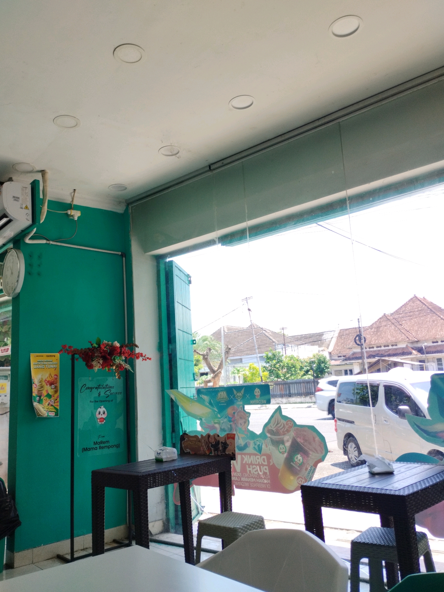 The image depicts the interior of a cafe, viewed from inside, looking out through a large window. The cafe has teal-colored walls and a white ceiling with recessed lighting fixtures. On the left wall, there's an air conditioning unit and a round clock. A floral arrangement in shades of red and yellow hangs on the wall. To the right of the flowers, a sign reads "Congratulations & Success" with text below in Indonesian.
In the foreground, two dark wicker tables are visible, one larger than the other. On the larger table, a pack of tissues is placed. Small, matching wicker stools are positioned around the tables. Outside the window, a street scene is visible with buildings, trees, and parked vehicles, including a white van. The general architecture and signage suggest a location in Indonesia, possibly Jember. The overall impression is that of a newly opened or recently decorated establishment. There are no people visible in the image.