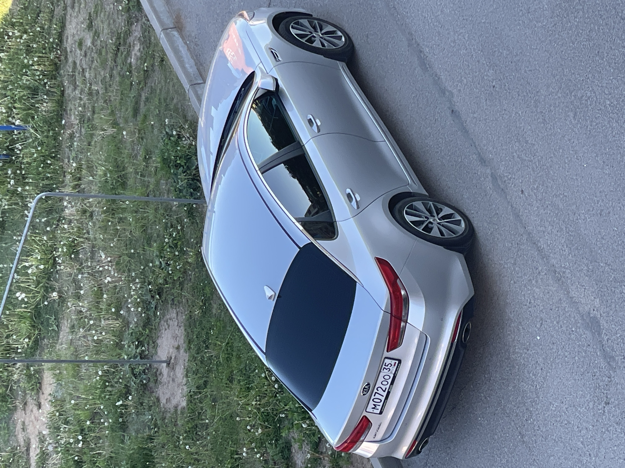 A silver Kia Optima sedan, model generation 2015-2020 (JF), is captured from a high-angle perspective, positioned parallel to a grey asphalt road. The vehicle displays Russian license plate M072OO 35, indicating registration in Vologda Oblast, Russia. The rear window is heavily tinted, and a white shark fin antenna is visible on the roof. Dual exhaust tips are integrated into the lower rear bumper. The car is equipped with multi-spoke alloy wheels. To the right of the vehicle, a grey concrete curb delineates the road from an unmaintained grassy area populated with numerous white dandelion seed heads. A thin, grey metal pole or railing extends upward from this grassy area on the left side of the frame. No individuals are present in the scene, and no specific actions are depicted. The lighting suggests daytime conditions. The exact location within the city of Vologda cannot be determined from the available visual information beyond the region code on the license plate.