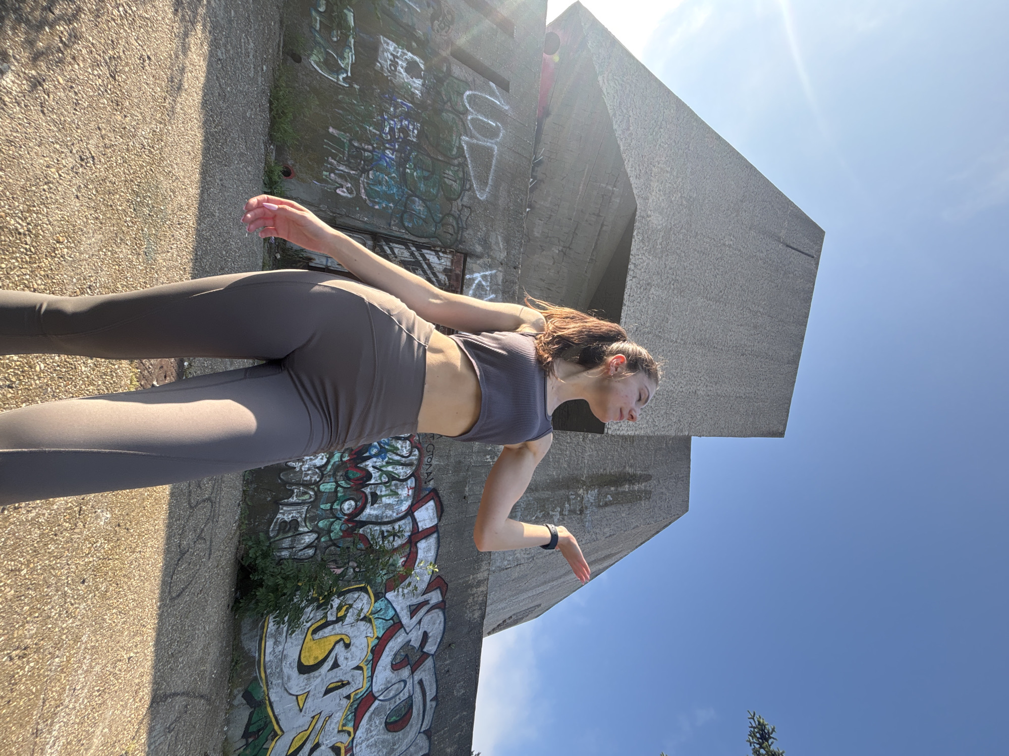 The image portrays a young woman in an athletic pose, set against a backdrop of monumental, graffiti-laden concrete structures under a brilliant blue sky. The photograph is oriented as if taken vertically but is displayed rotated, showing the woman seemingly moving horizontally across the frame.
The central subject, a woman with dark hair pulled back, is wearing dark grey or taupe athletic attire, consisting of high-waisted leggings and a ribbed crop top. She has a fitness tracker on her left wrist. Her expression is neutral, and her gaze is directed downwards, suggesting focus or contemplation. Her body is in a dynamic, mid-stride posture, with arms slightly bent and fingers curled.
The setting is characterized by imposing, brutalist-style concrete buildings that are extensively covered in colorful and intricate graffiti. These artworks feature stylized letters and tags in various hues, including yellow, white, green, and black. While most of the text is highly stylized and not easily decipherable into clear words, individual letters such as 'K', 'S', 'A', and 'R' can be discerned within the larger designs. The ground is a light-colored, textured pavement, possibly asphalt or concrete, with small patches of green vegetation growing near the base of the structures.
The bright sunlight, causing a visible lens flare in the upper left of the sky, indicates it is daytime, likely midday or early afternoon, with a clear, cloudless sky. The overall scene suggests an urban or industrial environment, consistent with the context of Varna, Bulgaria.