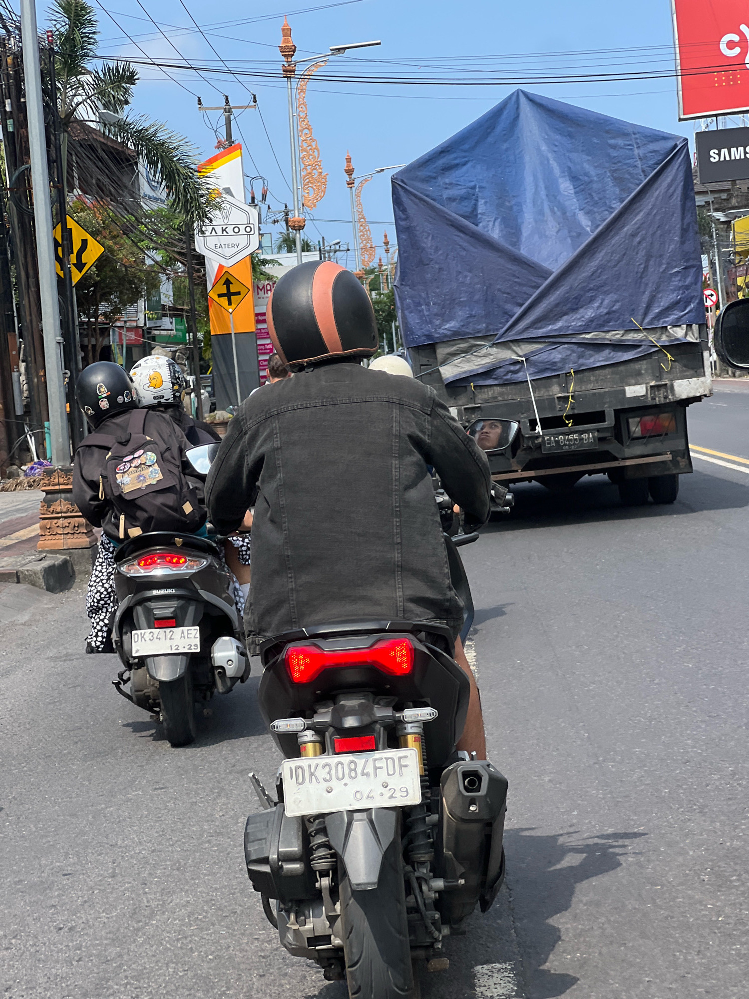 The image depicts a street scene in Denpasar, Indonesia, likely on a sunny day given the bright lighting and clear sky. The foreground is dominated by the rear view of two motorcycles traveling in tandem. The motorcycle on the right, closer to the camera, has a license plate visible that reads "DK3084FDF" with a date of "04-29". The rider is wearing a black denim jacket and a black and orange helmet. The motorcycle on the left, slightly further ahead, has a license plate reading "DK3412 AEZ" and a date of "12-29". The rider of this motorcycle is wearing a black jacket with a backpack adorned with several patches, and a black helmet. A passenger is seated behind this rider, also wearing a helmet, one white with a cartoon character and another black with pins.
In the background, a large truck covered with a blue tarp is moving in the same direction. The truck's license plate is visible and reads "EA-8455 BA". To the left of the road, there are utility poles with a dense network of wires, street lights with ornate decorations, and various signs, including a yellow directional sign with arrows and a sign for "LAKOO EATERY". Further in the background, buildings and more street signs are visible. The road surface is asphalt with painted lane markings. The overall impression is of everyday traffic on a busy street in a Southeast Asian city.