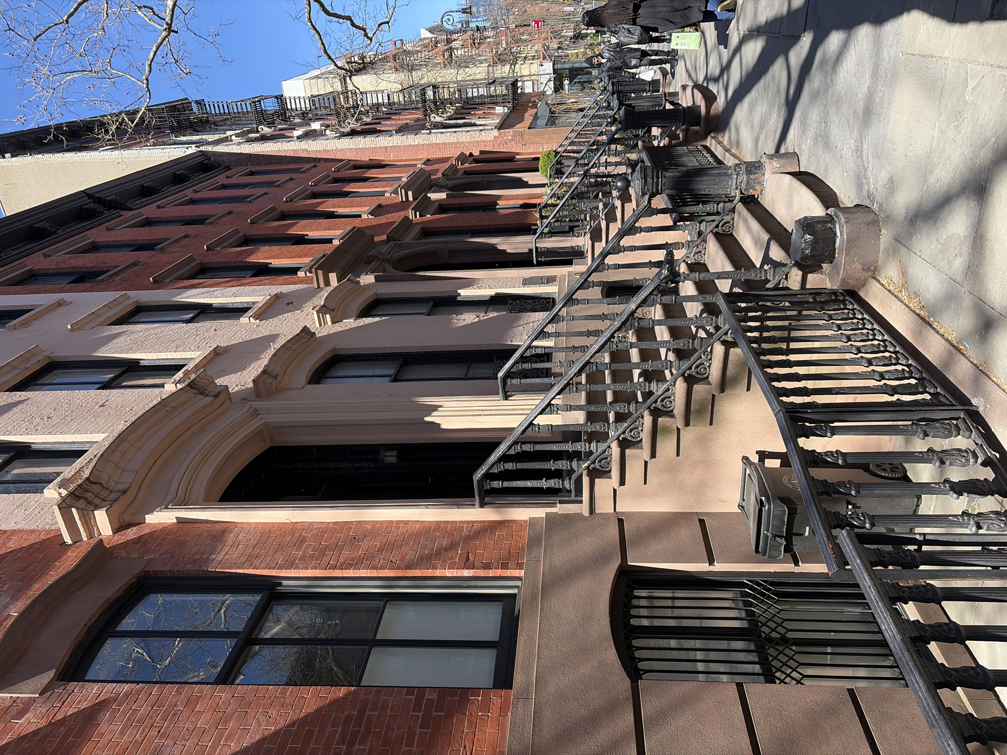 The image shows a street scene in New York City, United States. The focus is on a row of brownstone buildings with stoops and wrought-iron staircases leading up to the entrances. The buildings are constructed of red brick and beige stone. The windows are partially visible, and the details of the architectural trim and details of the buildings are visible. The sidewalk and a portion of the street are visible. Pedestrians are walking on the sidewalk. The image is taken during daylight hours, with shadows cast by the buildings and trees.