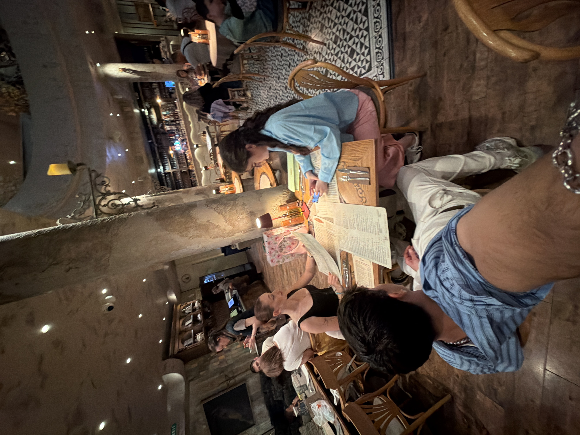 This image depicts an indoor scene at a restaurant or cafe in Sofia, Bulgaria, likely during the evening given the warm, ambient lighting. The setting features a rustic yet comfortable design with textured, light-colored walls, wooden tables and chairs, and decorative metalwork on the lighting fixtures.
In the mid-ground, a young woman with long dark hair, dressed in a light blue long-sleeved shirt and pink pants, is leaning over a table, seemingly interacting with items on it. To her right, a person is seated, wearing white pants and white sneakers, with another individual, visible from behind, wearing a blue striped shirt and a silver chain around their neck. Part of the photographer's arm, adorned with a silver chain, is visible in the bottom right corner.
Several other patrons are scattered throughout the background, seated at tables and engaged in conversation or looking at menus. A well-stocked bar area with bottles on shelves and various displays is noticeable in the distance. The floor shows a mix of wooden panels and a black and white patterned rug or tiled section. The overall atmosphere appears relaxed and inviting, with people enjoying their time in the establishment. Menus are visible on tables, but their specific text content is not legible.