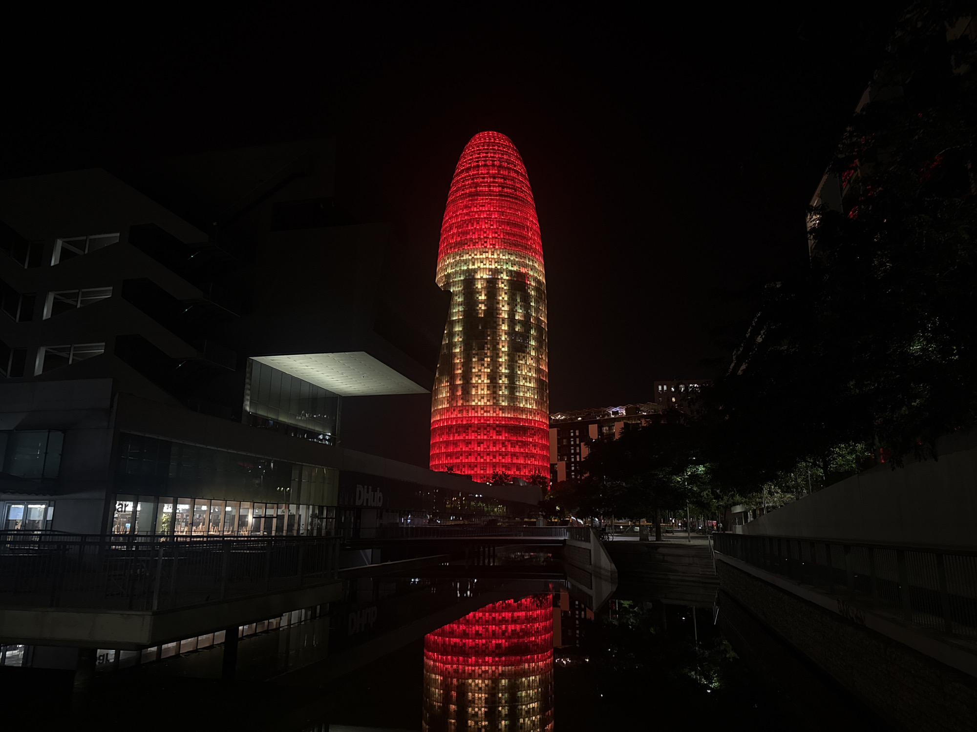 This is a nighttime photograph of the iconic Agbar Tower, also known as Torre Glòries, in Barcelona, Spain. The cylindrical skyscraper is illuminated with red and yellow lights, creating a striking visual against the dark night sky. The tower's distinctive shape, resembling a geyser or a bullet, is clearly visible.
In the foreground, a body of water, likely a canal or reflecting pool, mirrors the illuminated tower, doubling its impact. On the left side of the frame, a modern building with large windows and balconies is partially visible. The windows are lit from within, showing the interior of the building. A walkway with a railing runs in front of this building.
To the right of the tower, trees and other buildings are dimly visible in the background, indicating an urban environment. The overall scene is dark, with artificial lighting from the tower and surrounding buildings being the primary light sources. There are no people or vehicles directly visible in the frame. The composition emphasizes the architectural marvel of the Agbar Tower and its reflection.