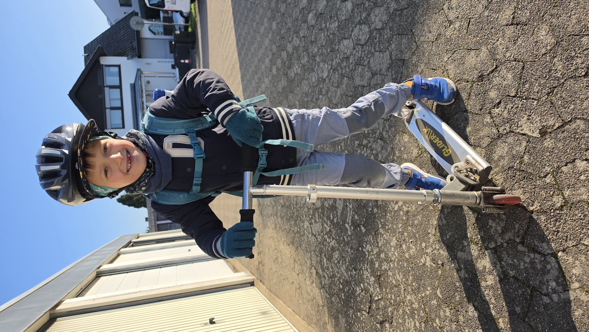 A young child wearing a helmet, jacket, backpack, gloves, and blue shoes is standing on a Gliderrik scooter on a paved surface. The child is smiling at the camera. Behind the child are buildings and a clear blue sky. The location is Unknown, Unknown.