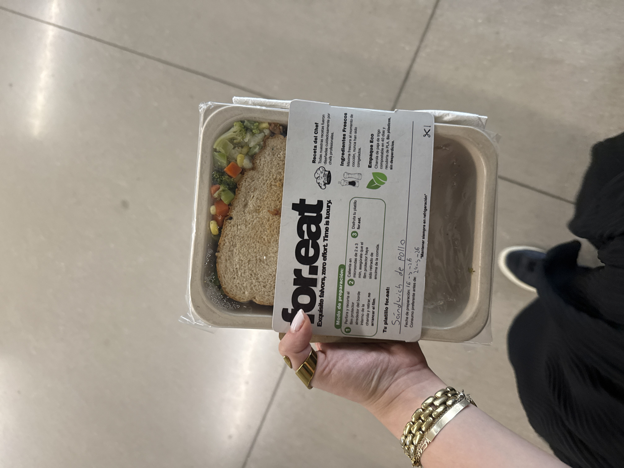 A person is holding a ready-to-eat meal in its container, likely indoors given the neutral-toned floor. The meal is a sandwich with a side of mixed vegetables, sealed in a clear plastic-covered tray. The tray has a white label with "for.eat" printed on it, along with cooking instructions and ingredient information in Spanish. The label also mentions the meal is a "Sandwich de pollo" (chicken sandwich). The production date on the label is 16-5-26, and the "consume before" date is 24-5-26. The person holding the meal is wearing a gold ring and a chunky gold bracelet. A portion of a black garment, possibly clothing, and the edge of a black shoe are visible in the background. There is no indication of this image being taken in Mexico City.