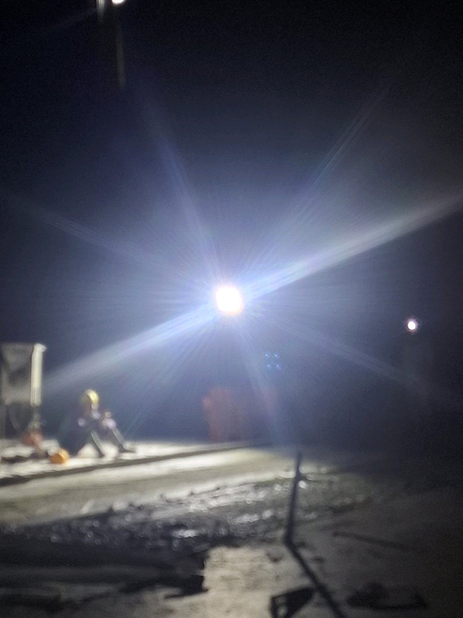 The image depicts a nighttime scene in Karangtengah, Indonesia, illuminated by a bright, intense light source creating a strong lens flare effect that dominates the center of the frame. In the foreground, the ground appears to be wet or reflective, with a dark, textured surface. To the left, a person wearing a yellow helmet is seated on the ground, with other figures and objects partially obscured by the darkness and glare. A vertical, light-colored structure is visible behind the seated figure. Further back, a faint orange object is present. The overall atmosphere is dark and industrial, with the bright light suggesting either artificial illumination for work or an event. The environment appears to be outdoors, possibly a street, construction site, or open area within the city.