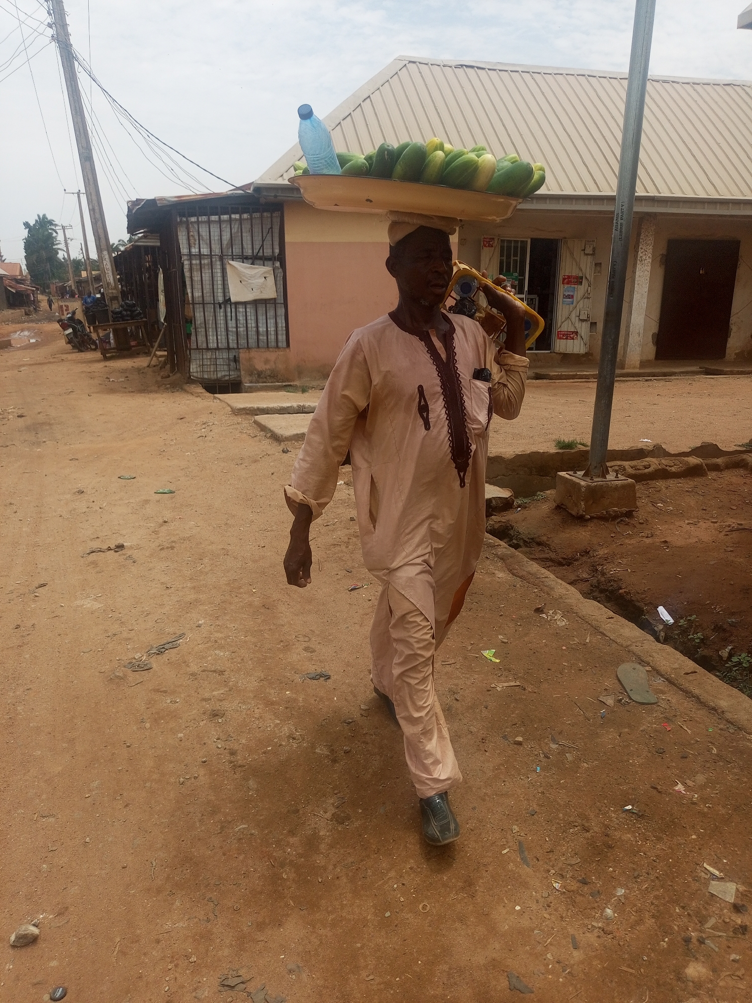 The image depicts a man walking down a dirt road in what appears to be Sabon Tasha, Nigeria. He is carrying a tray of what appears to be cucumbers and a plastic water bottle on his head. He is wearing a light brown tunic and pants, and black shoes. In his right hand, he is carrying what appears to be a yellow generator. The road is unpaved and has debris scattered on it. Buildings are visible in the background. The sky is overcast. A utility pole with wires is on the left of the frame.