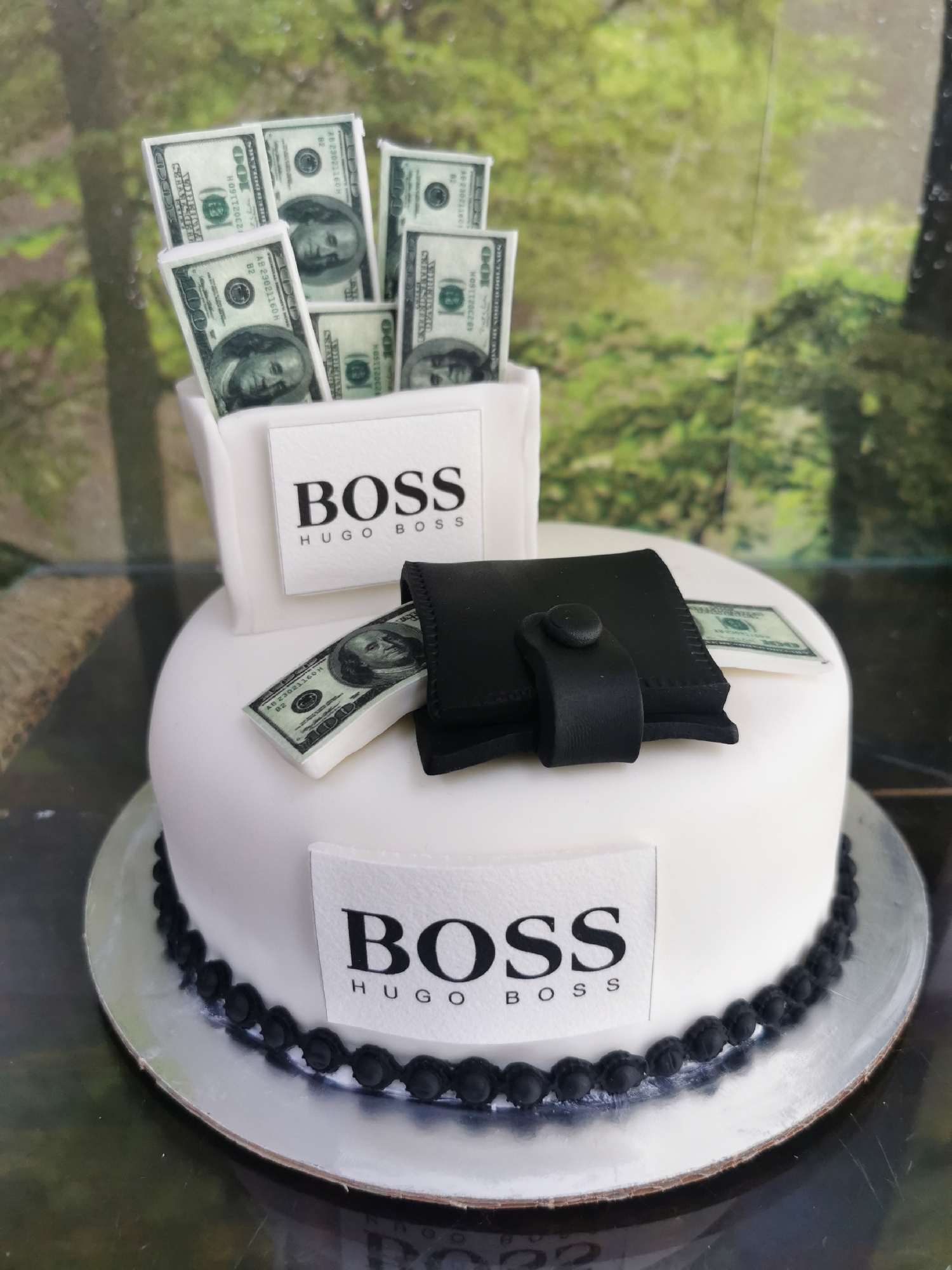 The image shows a cake decorated with elements symbolizing wealth and branding. A round cake with white frosting is centered on a metallic cake stand. The base of the cake is lined with a decorative trim of small, dark-colored spheres, possibly made of fondant. Two white square plaques are attached to the cake, each bearing the text "BOSS" in large, bold letters above the text "HUGO BOSS" in smaller letters. On top of the cake, a black wallet made of a fondant-like material is placed, with a couple of fondant $100 bills partially sticking out of it. Above the wallet, a white container is filled with more of these fondant $100 bills, creating a sense of abundance. The background appears to be a blurred outdoor scene with greenery. There is no indication in the image that the subject is in Varna, Bulgaria.
