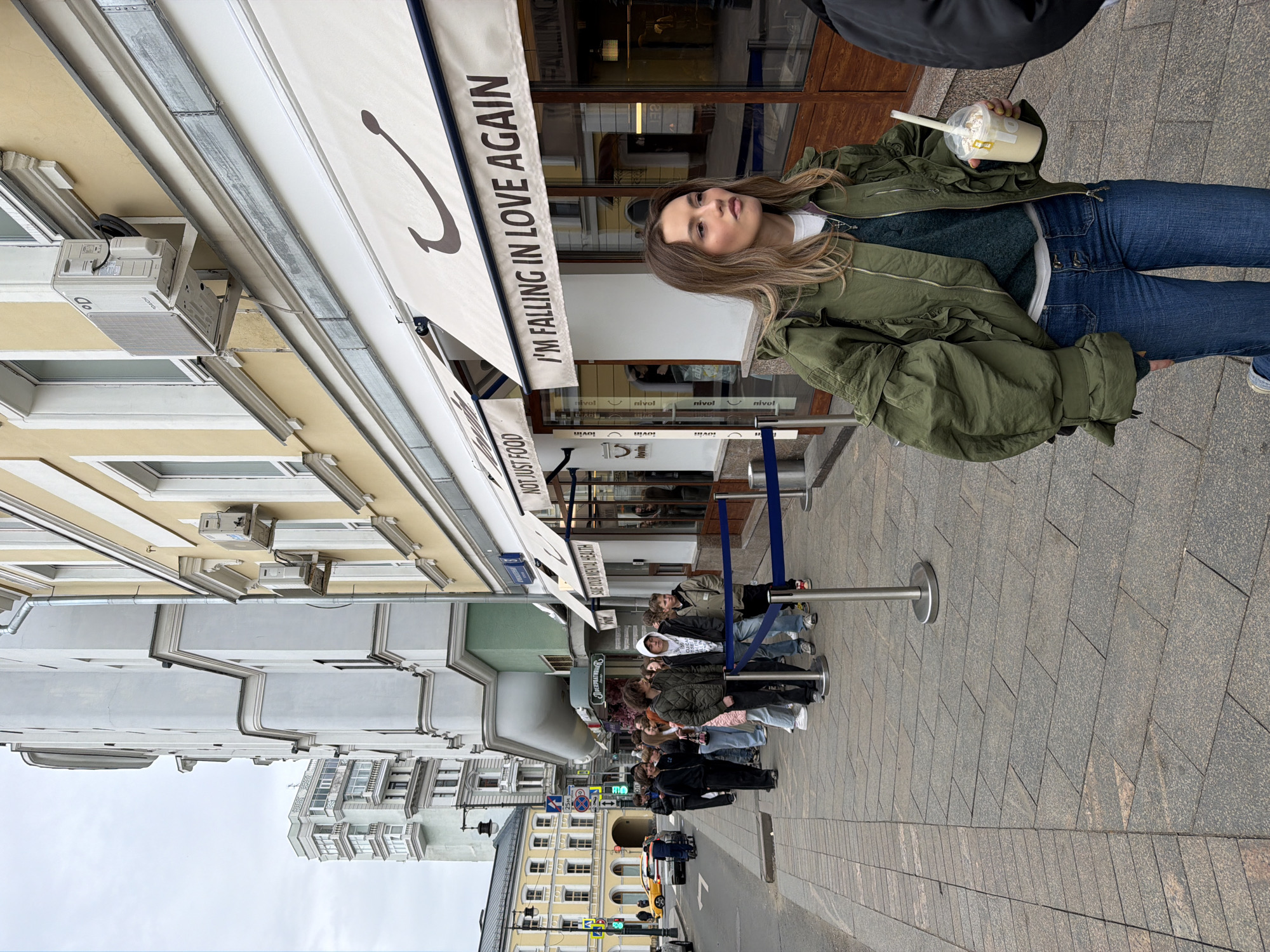 The image shows a young woman standing outside a cafe in Moscow, Russia. She is holding a beverage in a plastic cup and is wearing a green jacket and blue jeans. The cafe is decorated with an awning that reads "I'm falling in love again." There is a queue of people in the background. The street has old buildings on one side and modern structures on the other. There is a traffic light and some cars in the distance.