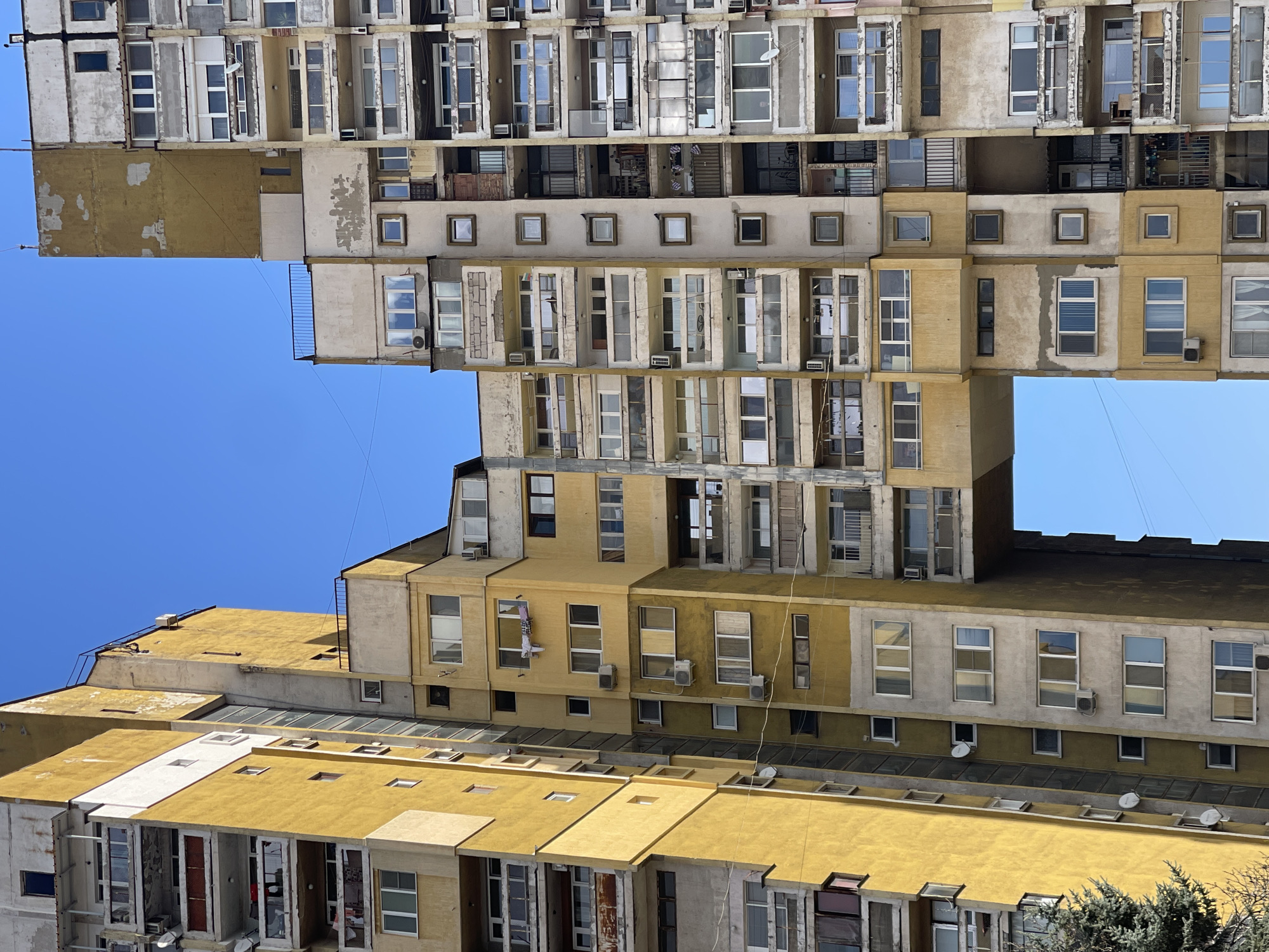 The image depicts a high-angle, rotated view of an apartment building complex in Sofia, Bulgaria, set against a clear blue sky. The architecture is characterized by stacked and offset sections, creating a layered effect. The buildings are predominantly yellow and beige with numerous windows and some air conditioning units. There are no people visible. The environment consists of urban buildings and blue sky.