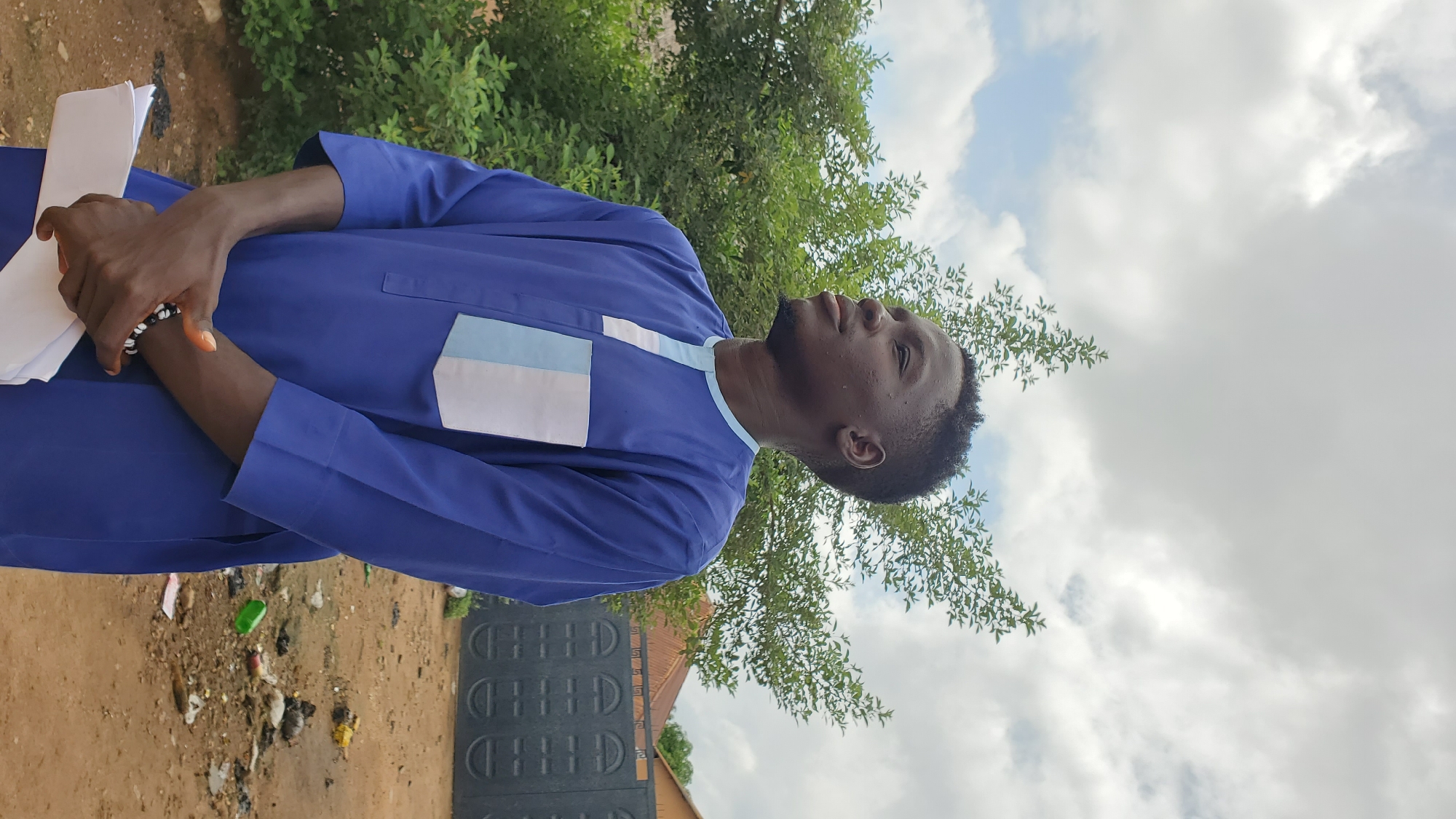 A young Black male stands outdoors, facing right and looking upwards. He is wearing a blue garment with light blue and white accents on the chest. His hands are clasped in front of him, holding a few white papers. A bracelet with black and white beads adorns his left wrist. Behind him, a dense green tree is visible, with the suggestion of buildings and a dark metal gate in the background. The ground in front of him is dirt, with some scattered debris. The sky is overcast with white and gray clouds. The scene appears to be in the city of Jalingo, Nigeria.
