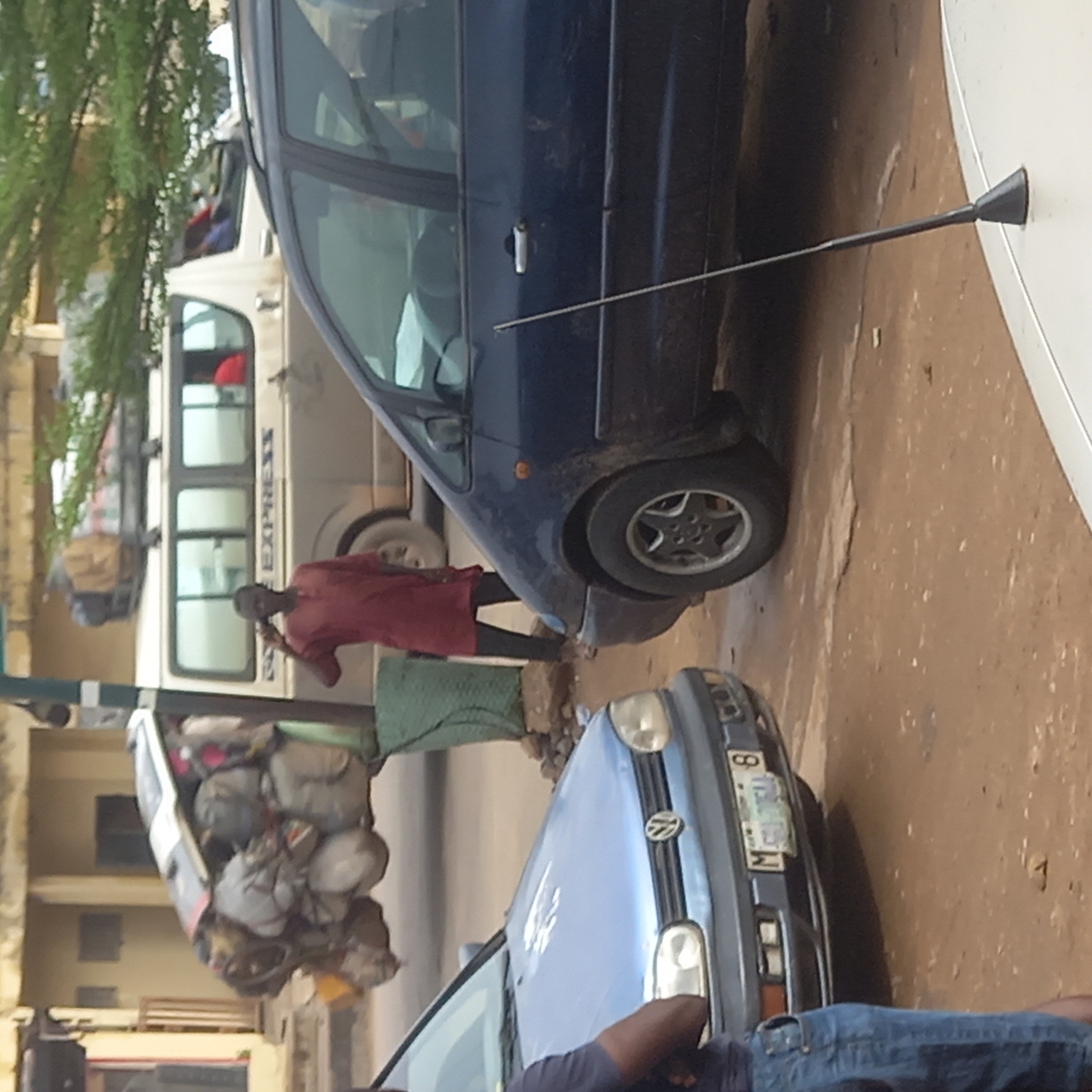 In Dadi Riba, Nigeria, the image shows a busy street scene with several vehicles present. A dark blue minivan is parked on the right side of the image. A light blue Volkswagen Golf is parked further down the street. A white "EXPRESS" van, heavily loaded with luggage on its roof, is visible behind the blue minivan. A person wearing a red shirt and dark pants is standing next to the "EXPRESS" van. A person wearing jeans is visible at the bottom of the image, near the blue Volkswagen Golf. A green cloth hangs near the minivan. A satellite dish is mounted on a wall on the right.