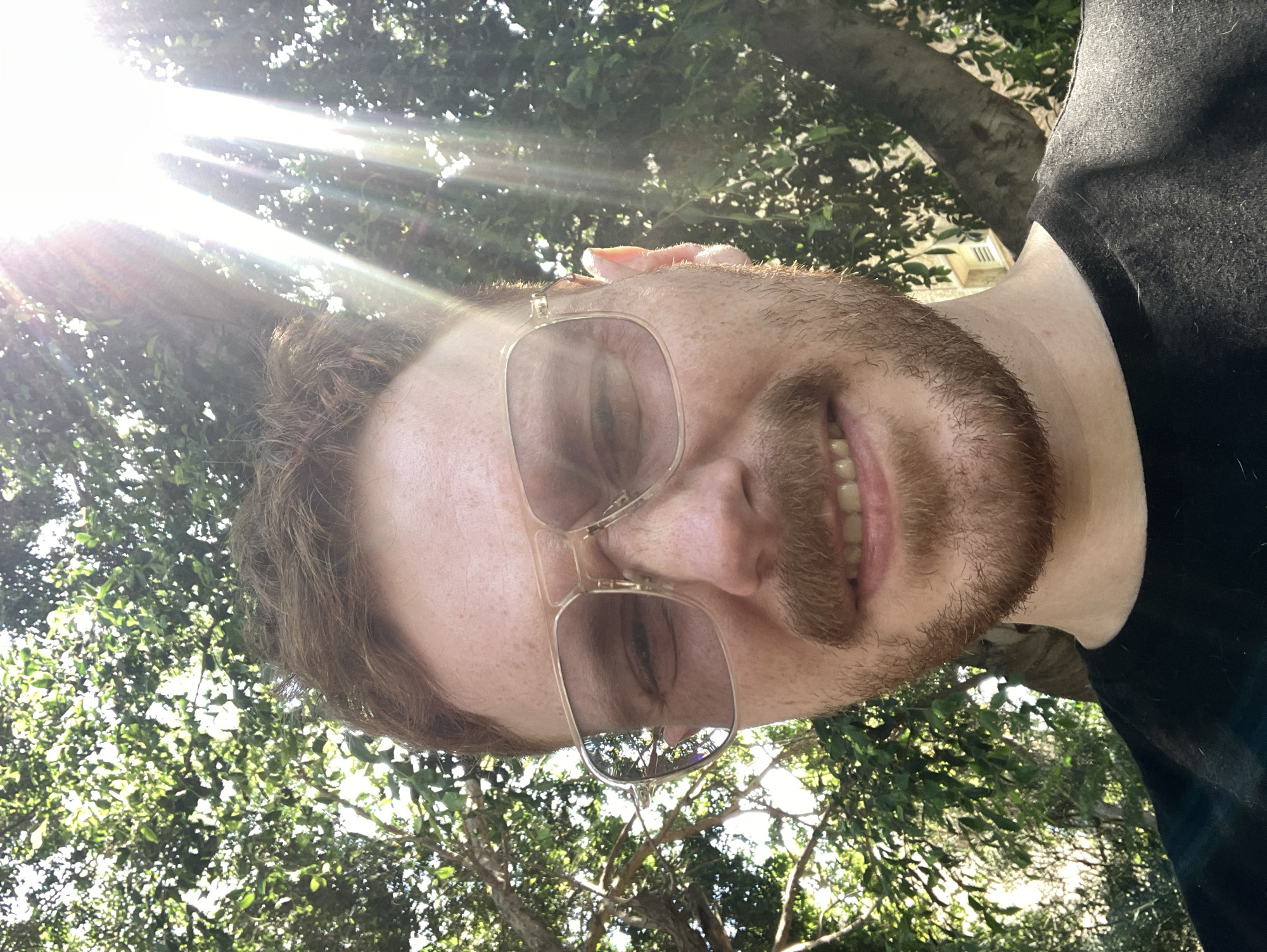 A man with fair skin, freckles, and reddish-brown hair is pictured in a selfie taken outdoors, with sunlight streaming through trees. He is wearing large, square-framed glasses with a light-colored tint, and has a mustache and beard. His mouth is slightly open, revealing his teeth. He is wearing a dark t-shirt. The background consists of lush green foliage and tree trunks, with a hint of a building visible in the upper right portion of the frame. The lighting suggests a sunny day. The overall scene implies a casual outdoor setting, possibly a park or garden, within Tel Aviv, Israel.