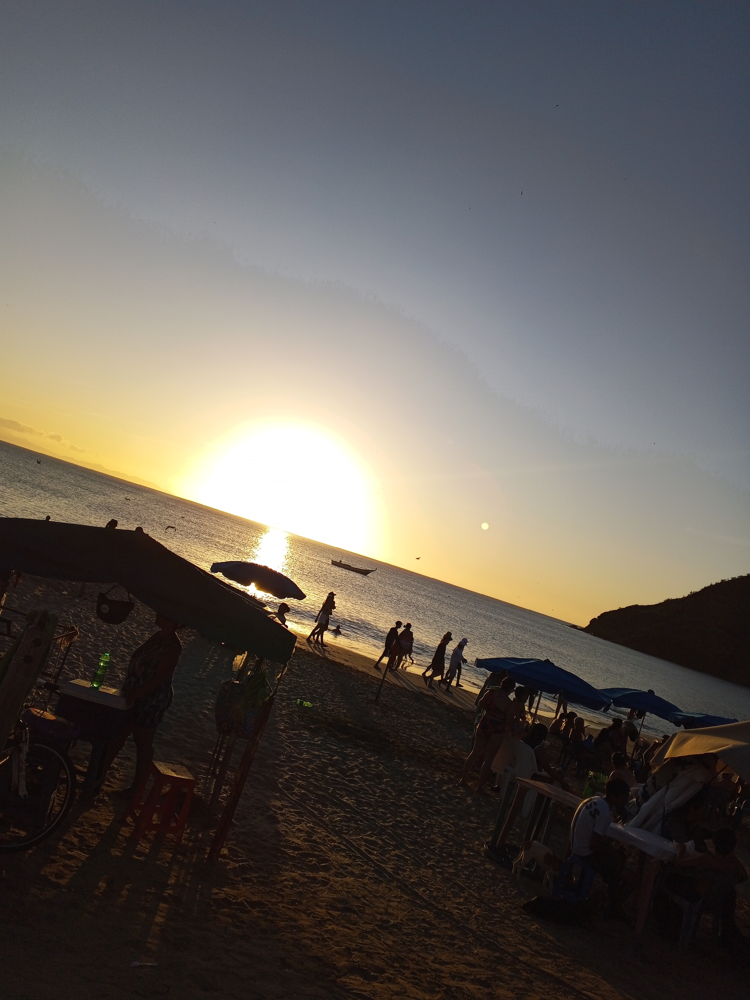 Captured at sundown in Juan Griego, Venezuela, the image portrays a vibrant beach scene. The setting sun casts a golden hue over the ocean, where a small boat is visible in the distance. A group of people stroll along the shoreline, while others are gathered near tables and beneath umbrellas, suggesting a lively beachside atmosphere. The foreground features market stalls, offering services and food. The scene is dynamic, portraying people enjoying an ocean view at sunset, with stalls providing food and other services.