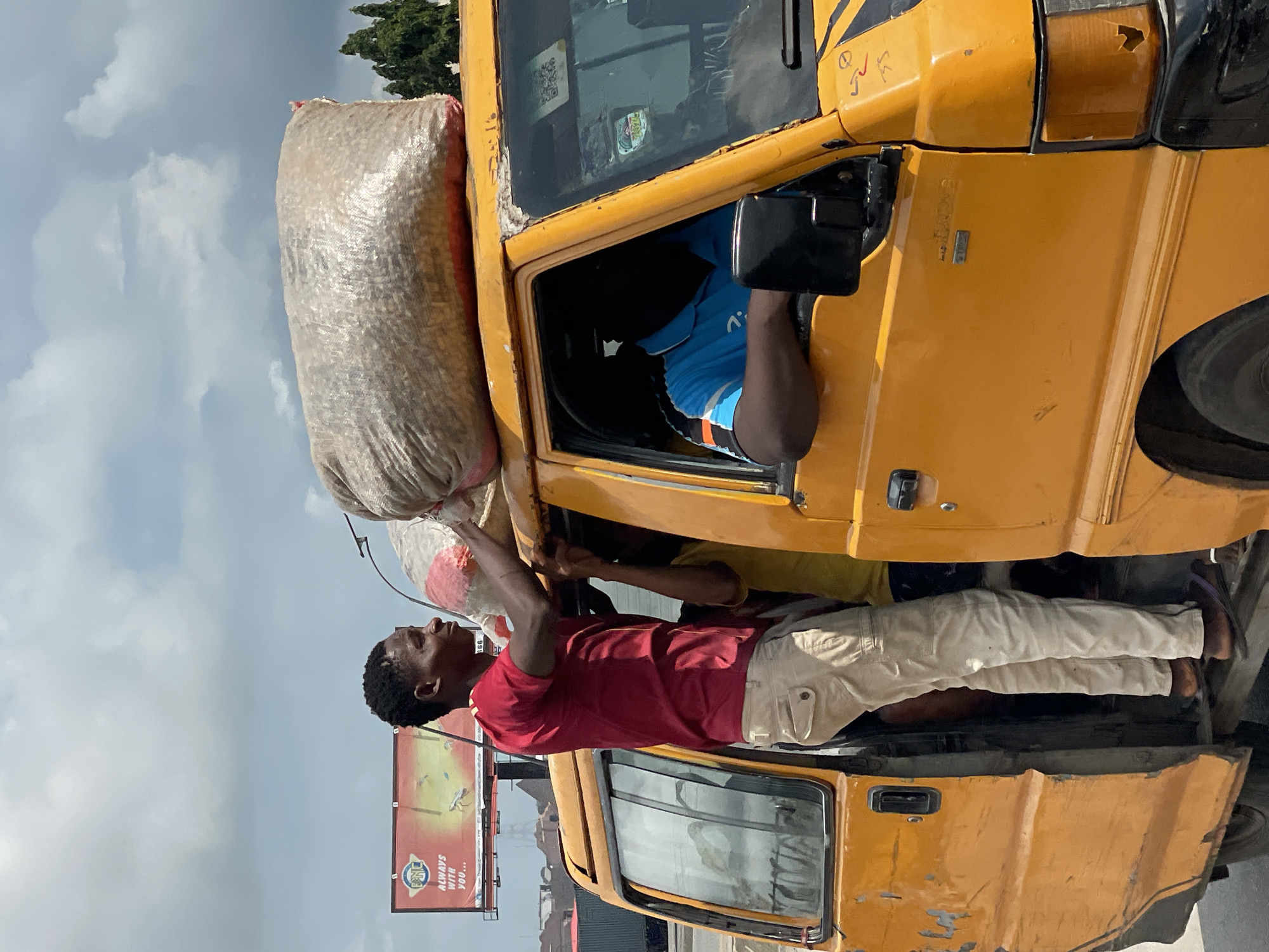 In Fegge, Nigeria, a yellow minibus is being loaded with large sacks of goods on its roof. A young Black man, wearing a red t-shirt and khaki pants, stands on the running board of the open driver's side door, reaching up to adjust a bulky, light-colored sack. His head is tilted back, and he appears to be straining slightly. Behind him, another Black person in a yellow shirt is visible inside the bus, partially obscured. The driver, a Black man wearing a blue collared shirt, is seated behind the steering wheel, looking forward. The minibus shows signs of wear, with peeling paint and scuffs on its exterior. In the background, a billboard with red and yellow lettering is visible, along with trees and a cloudy sky. The scene captures a moment of commerce and transportation in an urban setting.