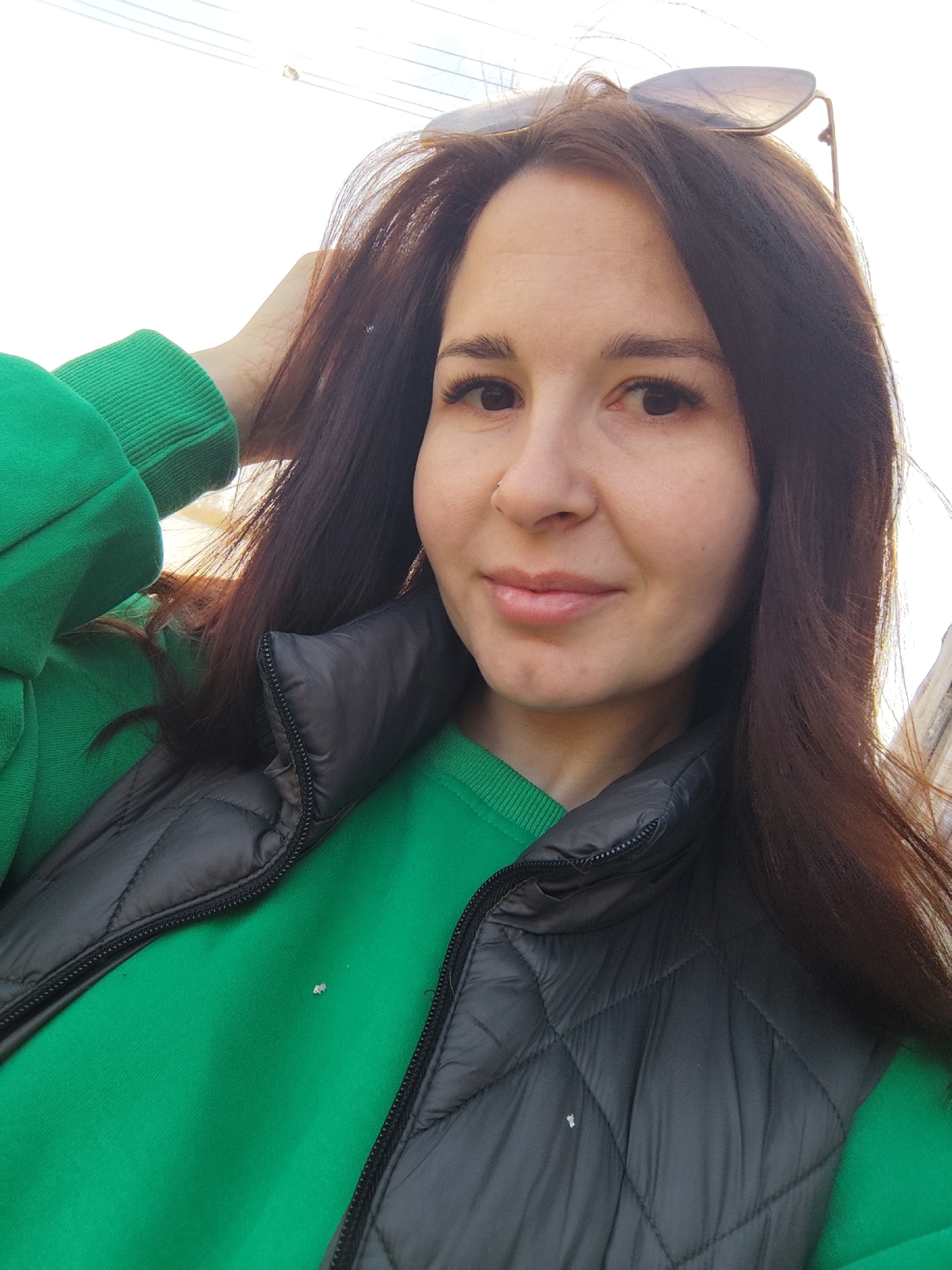 A young woman is posing for a selfie, outdoors, with her hand running through her brown hair. She is wearing sunglasses perched on her head, a green hooded sweatshirt, and a black quilted vest. The location is possibly in the city of Фоки, Russia, given the prompt instructions. The background is bright, with some blurry shapes possibly indicating urban elements. The light suggests it is daytime. A small white piece of debris is visible on the green sweatshirt.