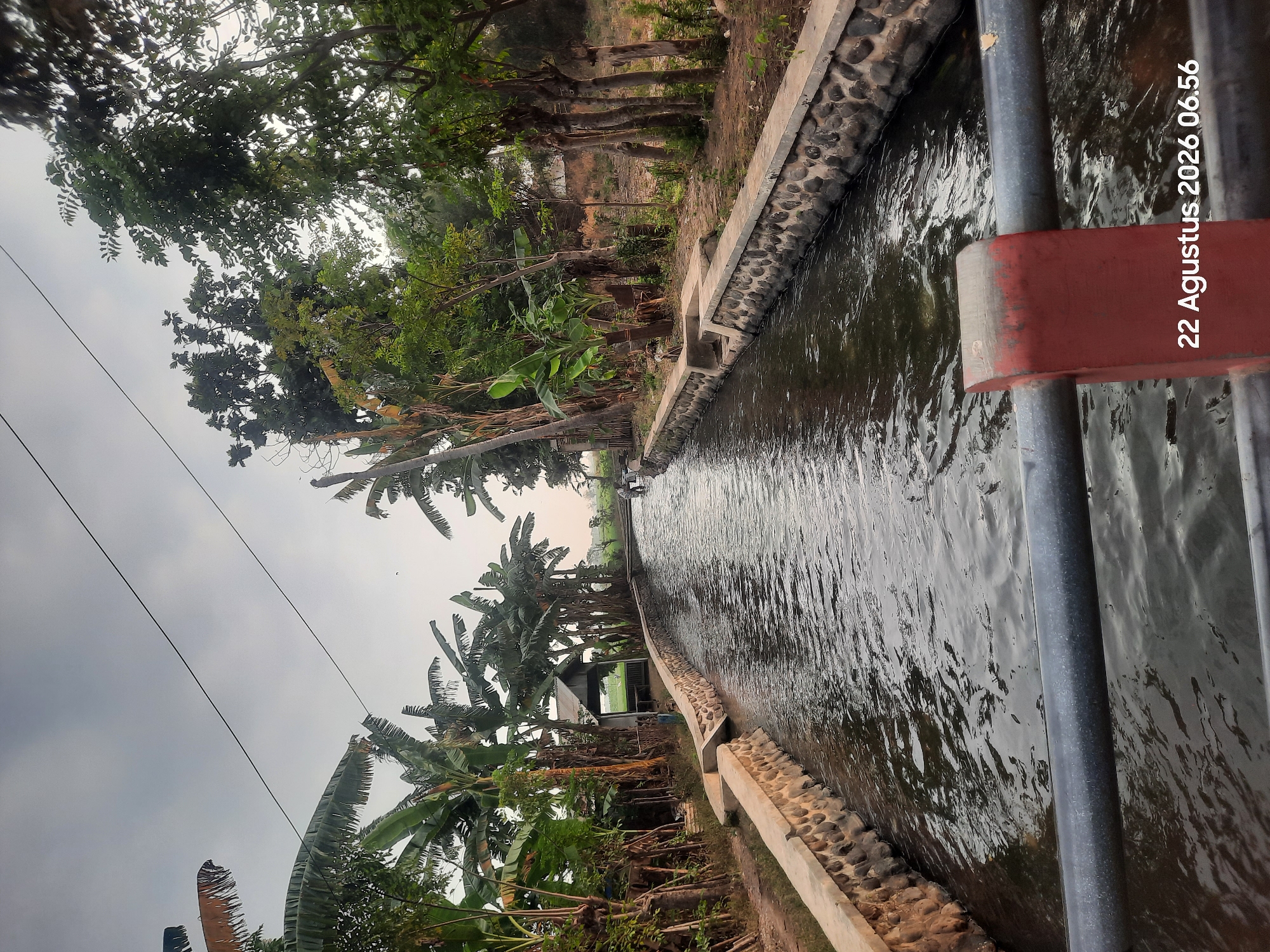 The image shows a canal running through a rural area, likely Karangbong, Indonesia, based on the overlaid text. The canal is flanked by stone and concrete walls, with lush vegetation, including banana trees, on either side. In the distance, a small building with a green roof is visible, and a few people can be seen near the canal. The water is calm, reflecting the overcast sky. The foreground features a metal railing with a red support structure, and the date "22 Agustus 2026 06.56" is superimposed on the image.