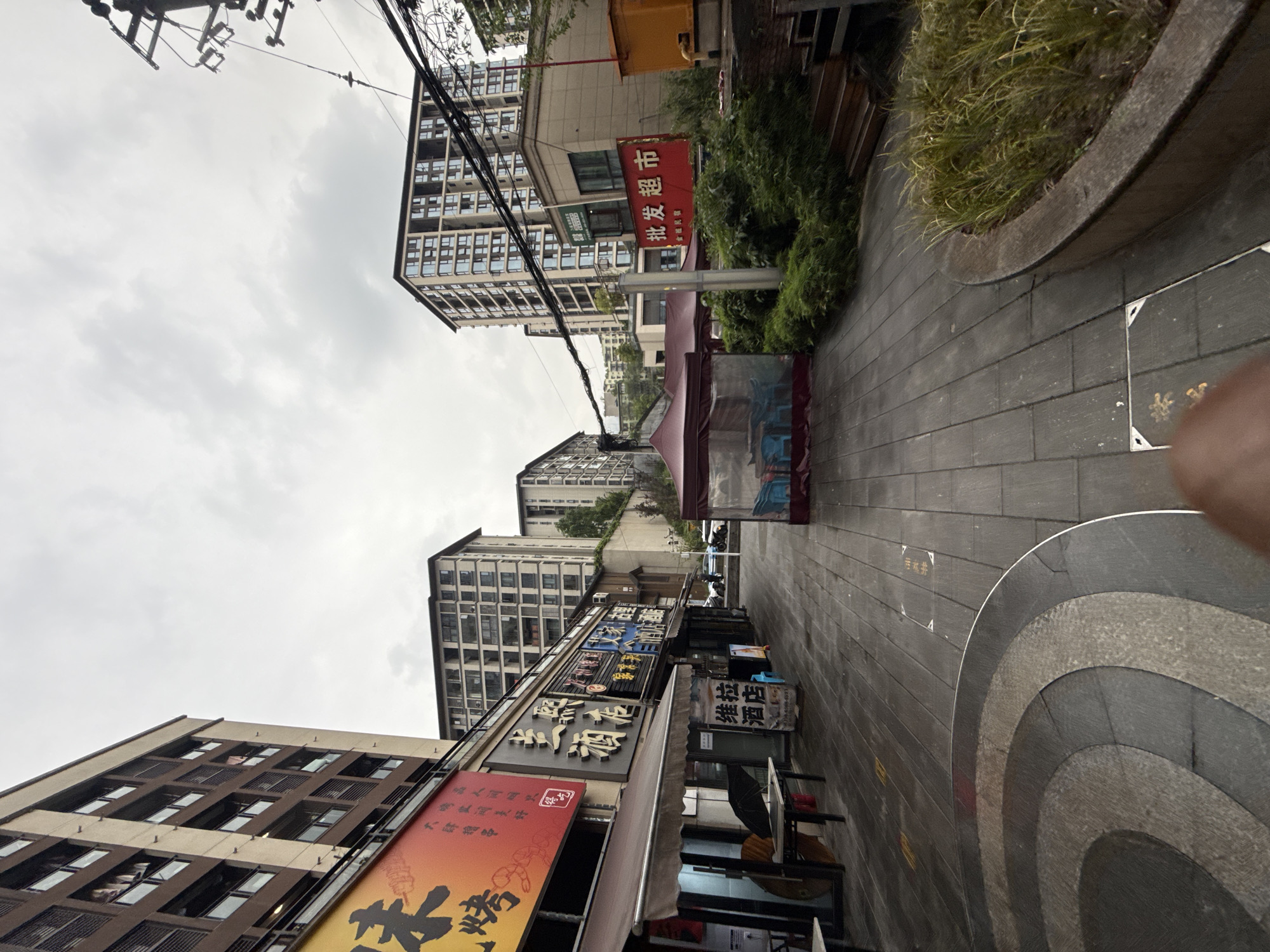 Here's a description of the image:
**Scene Overview:** The image shows a street scene in Shapingba District, China, characterized by a mix of commercial and residential buildings. The shot is taken at a slightly tilted angle, looking down a street lined with shops and tall apartment buildings. The sky is overcast.
**Subjects and Objects:**
* **Buildings:** The scene is dominated by high-rise apartment buildings and various commercial structures. These appear to be modern with many windows.
* **Shops:** The street-level shops have awnings and storefront signage with Chinese characters. The signs indicate various businesses including a supermarket, restaurants, and likely other retail establishments.
* **Street:** The paved street has a circular design element in the pavement.
* **Signage:** There are numerous signs in Chinese characters, including one for a "Supermarket" and advertisements for restaurants.
* **Utility Infrastructure:** Wires and utility poles are visible, common elements in many urban scenes.
**Environment:** The setting is an urban area in China. The time of day appears to be daytime, given the level of light despite the cloudy sky.