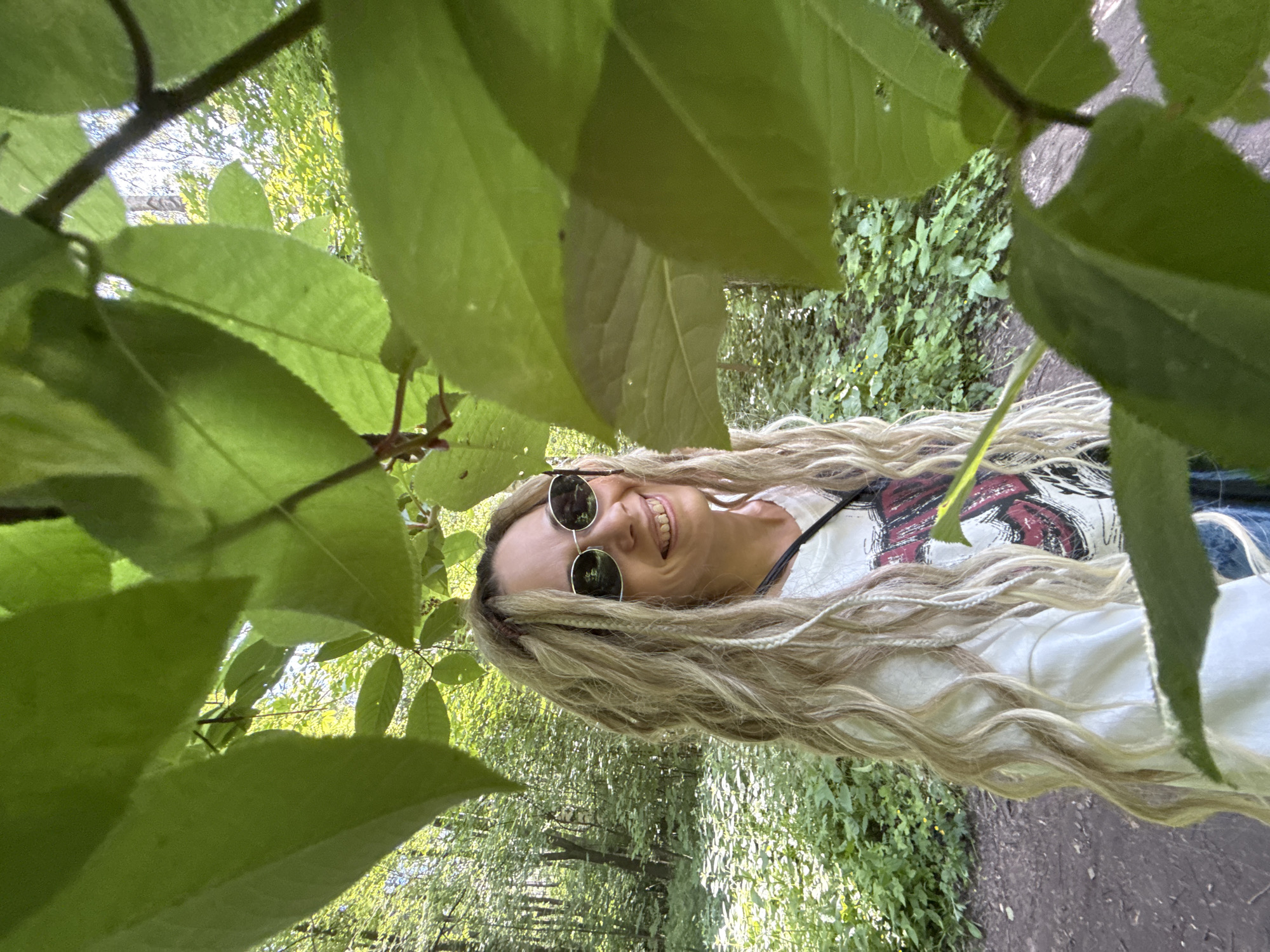 The image captures an adult female outdoors in a densely vegetated area, identified as Rusanovo, Russia. She is positioned slightly right of the frame's center, facing and smiling towards the viewer. Her long, blonde, wavy hair contains multiple lighter blonde dreadlocks or braided extensions, some secured with a reddish band on her left side. She wears round, dark-lensed sunglasses and a white t-shirt featuring a partially visible red and black graphic design on the chest. A dark strap is visible over her right shoulder. The immediate foreground is heavily framed by large, vibrant green leaves and branches. The background reveals a sunlit forest environment with numerous green trees, including a birch-like trunk, and dense undergrowth. A dirt path is visible on the ground to the left of the subject. Natural daylight illuminates the scene.