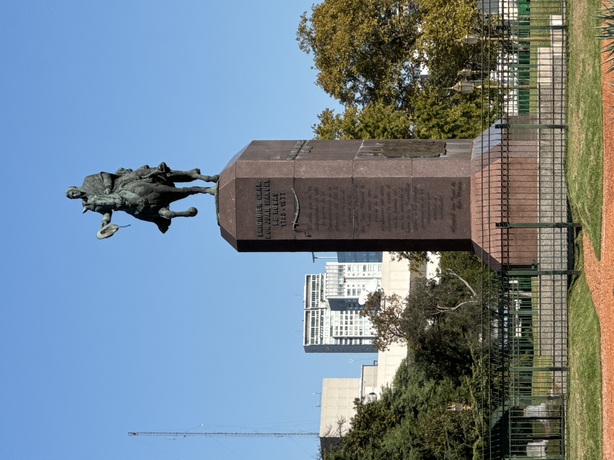 The image shows the monument to Brigadier General Don Juan Manuel de Rosas in Buenos Aires, Argentina. It includes the bronze equestrian statue of Rosas atop a tall, dark stone pedestal with text etched into its sides. The monument is surrounded by a fence, with trees and buildings visible in the background under a clear blue sky. A construction crane is also visible in the distance.