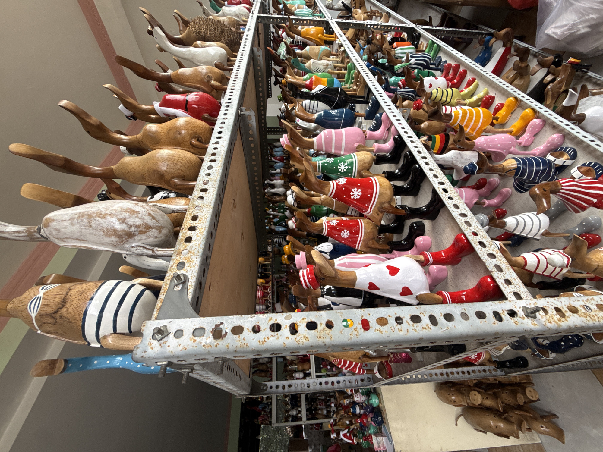 The image displays a retail setting, likely a souvenir shop, featuring a large collection of wooden duck figurines. These figurines are diverse in their attire, with many dressed in colorful outfits, including dresses, sweaters, and boots. Some ducks wear hats, and others have patterned designs on their bodies. The ducks are displayed on multiple shelves of a metal rack. In the background, more shelves are visible, also filled with similar figurines and other decorative items, suggesting a bustling marketplace or workshop. The setting appears to be indoors, with natural and artificial lighting illuminating the scene. The overall impression is one of a vibrant and abundant display of handcrafted goods.