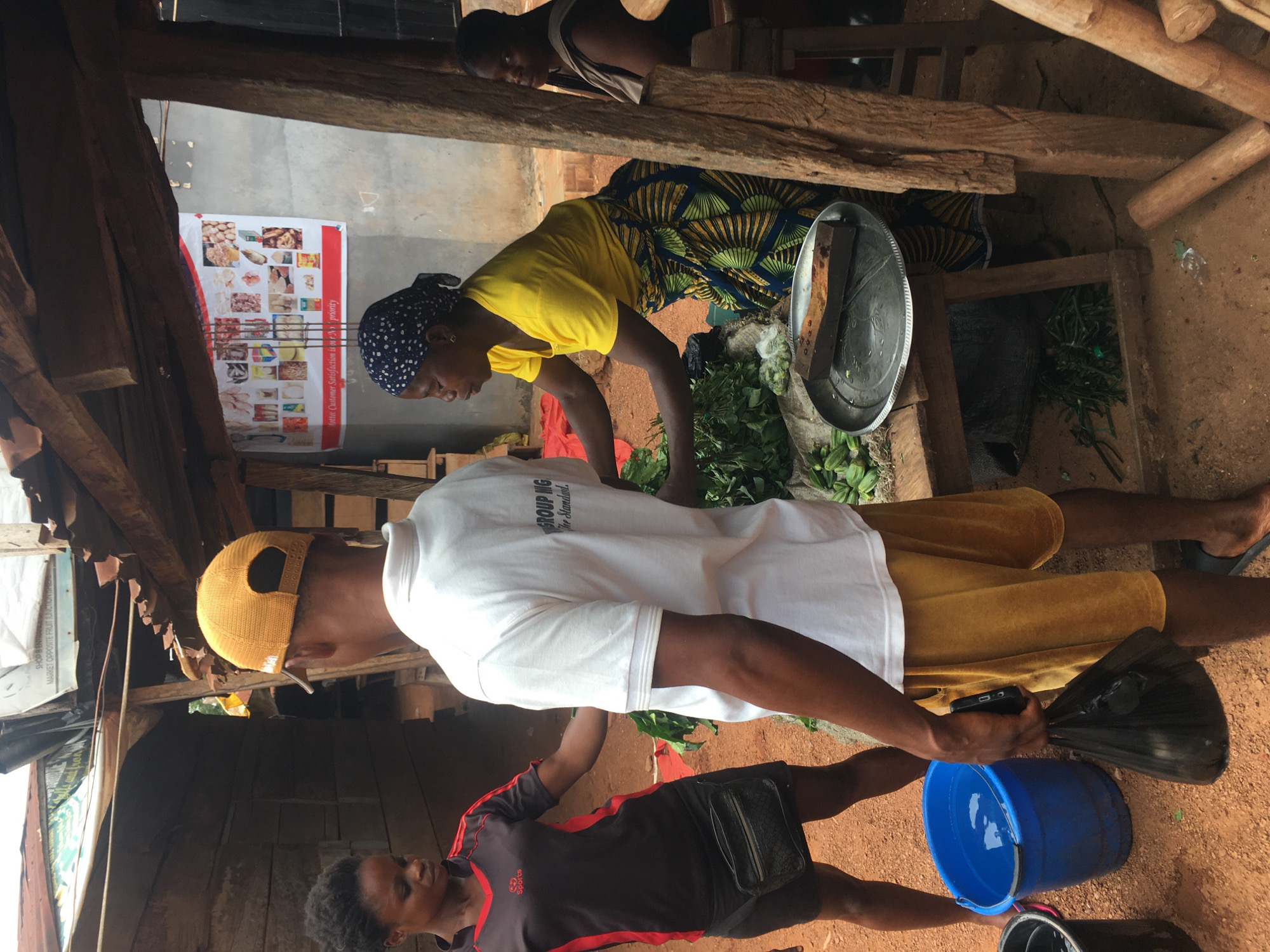 This vibrant image depicts a bustling outdoor market stall in Enyibichiri, Nigeria, under natural daylight. The setting is characterized by a rustic, open-air structure constructed from rough-hewn wooden beams and posts, with a dirt ground.
In the mid-ground, a woman wearing a bright yellow top, a traditional patterned wrapper, and a blue patterned headscarf is actively engaged in sorting or preparing large piles of fresh green leafy vegetables on a low counter or table. Beside the greens, a silver tray with a dark, rectangular object, possibly a weight, rests on the counter. Behind her, partially obscured by wooden beams, another person in a light-colored top is visible.
In the foreground, a man wearing a white t-shirt, a yellow baseball cap, and yellow shorts stands, holding a black plastic bag and a mobile phone. Near his feet, a blue plastic bucket, partially filled with water, sits on the ground. Another individual, wearing a black and red "Sports" branded top, is partially visible near the man, facing the bucket.
A white poster is affixed to a wall, showcasing a variety of food items, including meats, fish, and baked goods. Visible text on the poster reads "lotto: Customer Satisfaction is on priority," "SHOP B ENTRA," and "MARKET OPPOSITE FRUIT JUNCTIONISHIER." Other text fragments like "GROUP NIG" and "The Standard." are also discernable. The scene captures a moment of daily commerce and interaction within a local marketplace.