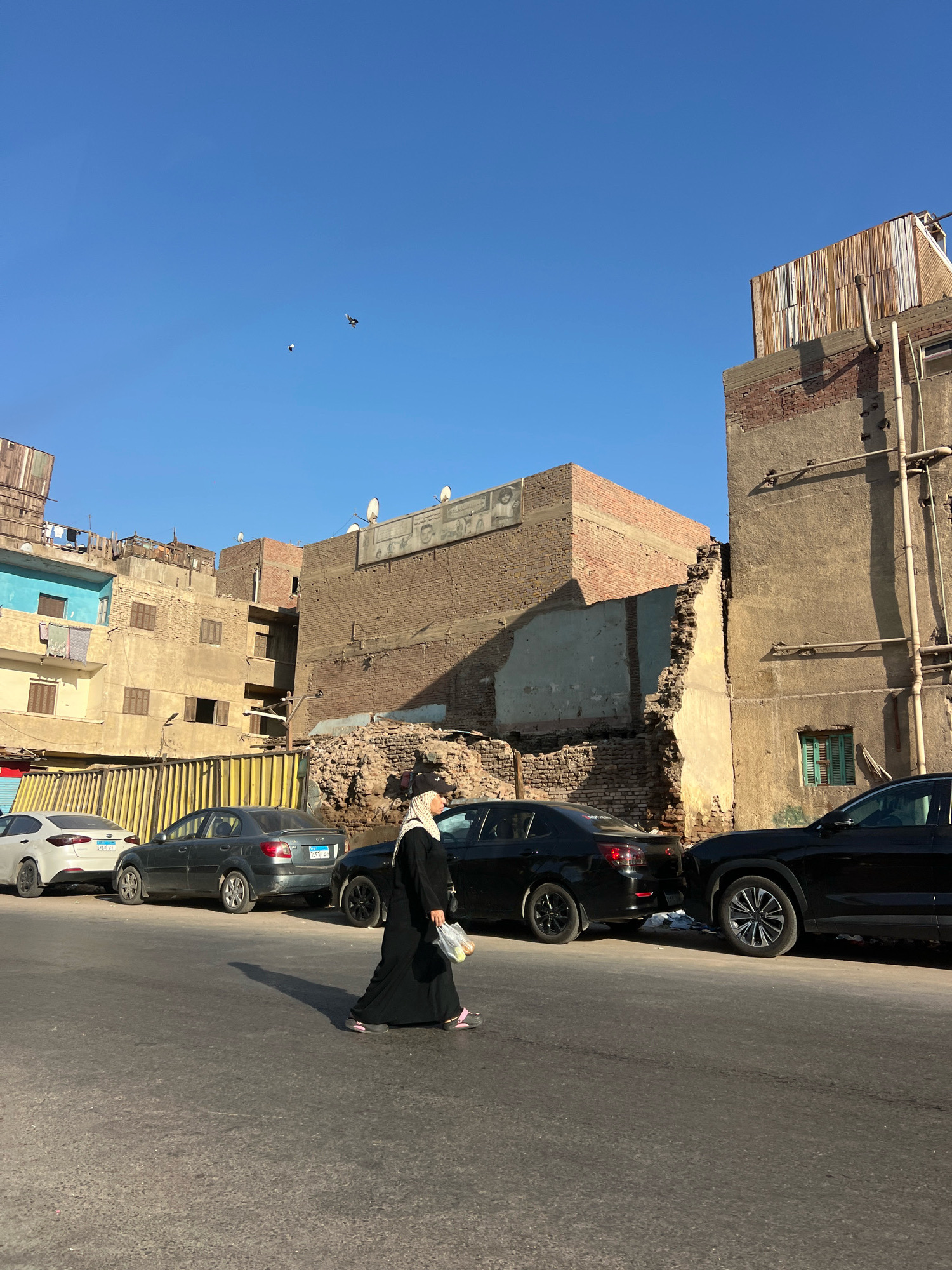 A woman in a black abaya and hijab walks across a street in Giza, Egypt, casting a long shadow in the late afternoon sun. She carries a plastic bag filled with what appear to be fruits. Several cars are parked on the street, including a white Kia, a dark gray sedan, and two black sedans. Behind the cars, a row of buildings displays various states of repair, with some appearing weathered and others showing signs of construction or demolition. One building has a large section of its brick wall exposed, revealing structural damage. Above the buildings, a clear blue sky is visible, with two birds in flight. The scene suggests a typical urban environment with residential and commercial structures.