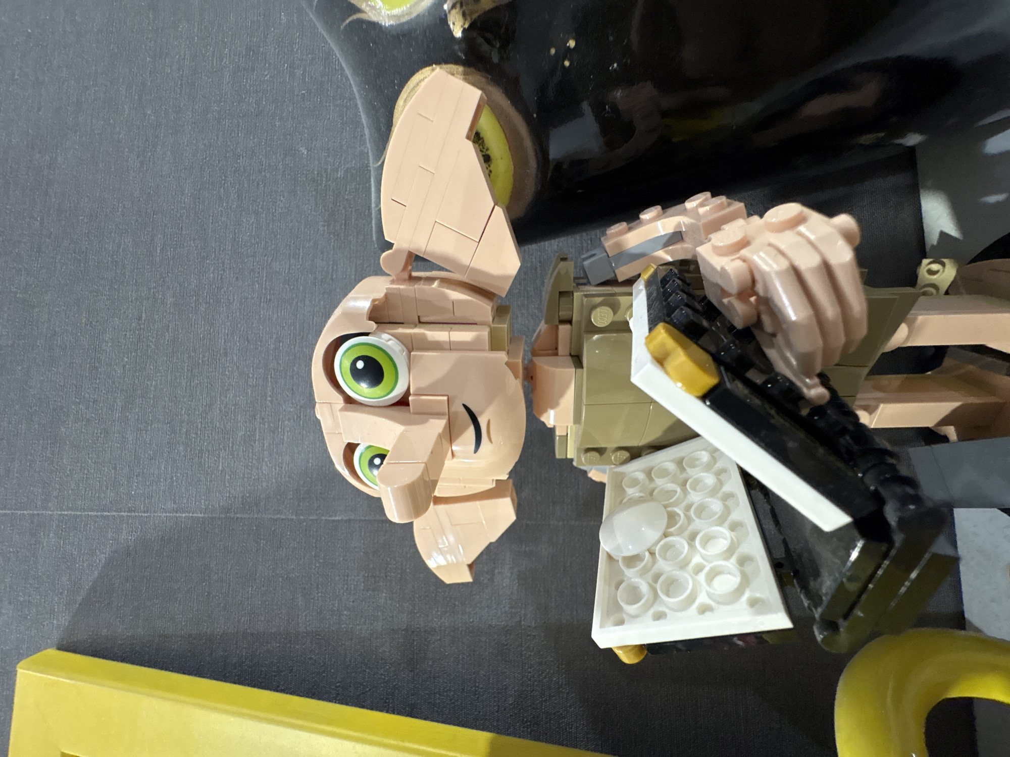 A close-up, slightly rotated image shows a Lego figurine resembling Dobby from the Harry Potter series. The figure is composed of various Lego bricks and is mostly beige with green eyes. It is holding what appears to be a book with a white cover, white details, and a black spine. A portion of a yellow object is visible at the bottom of the frame. The background is a plain gray surface.